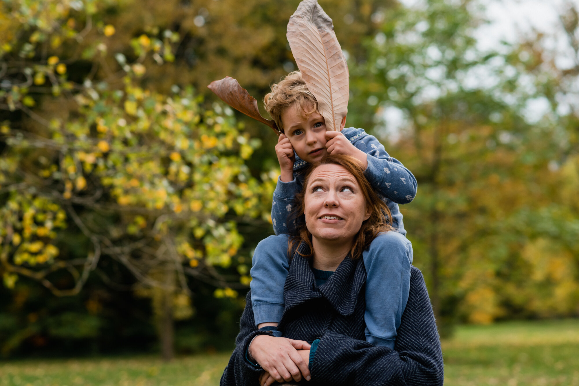  A boy in a blue top and trousers is seen putting two big oval shaped leaves by his ears, as he sits on his mother's shoulders and she is looking up at him, in Kew Botanical Gardens, Surrey. 