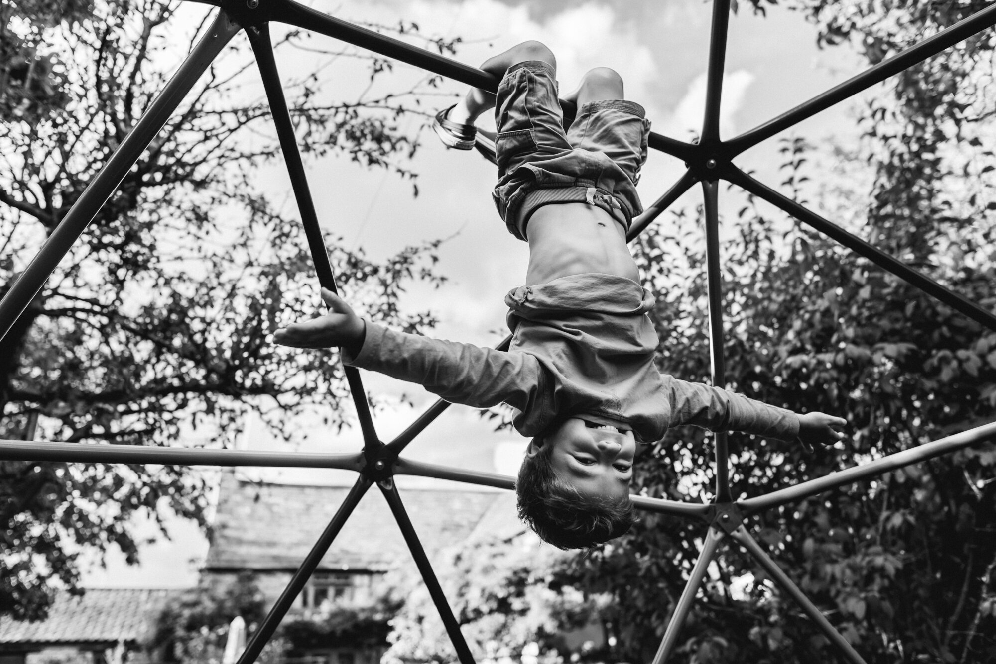  Black and white photograph of a child hanging upside-down on a metal dome climbing frame in the garden. 