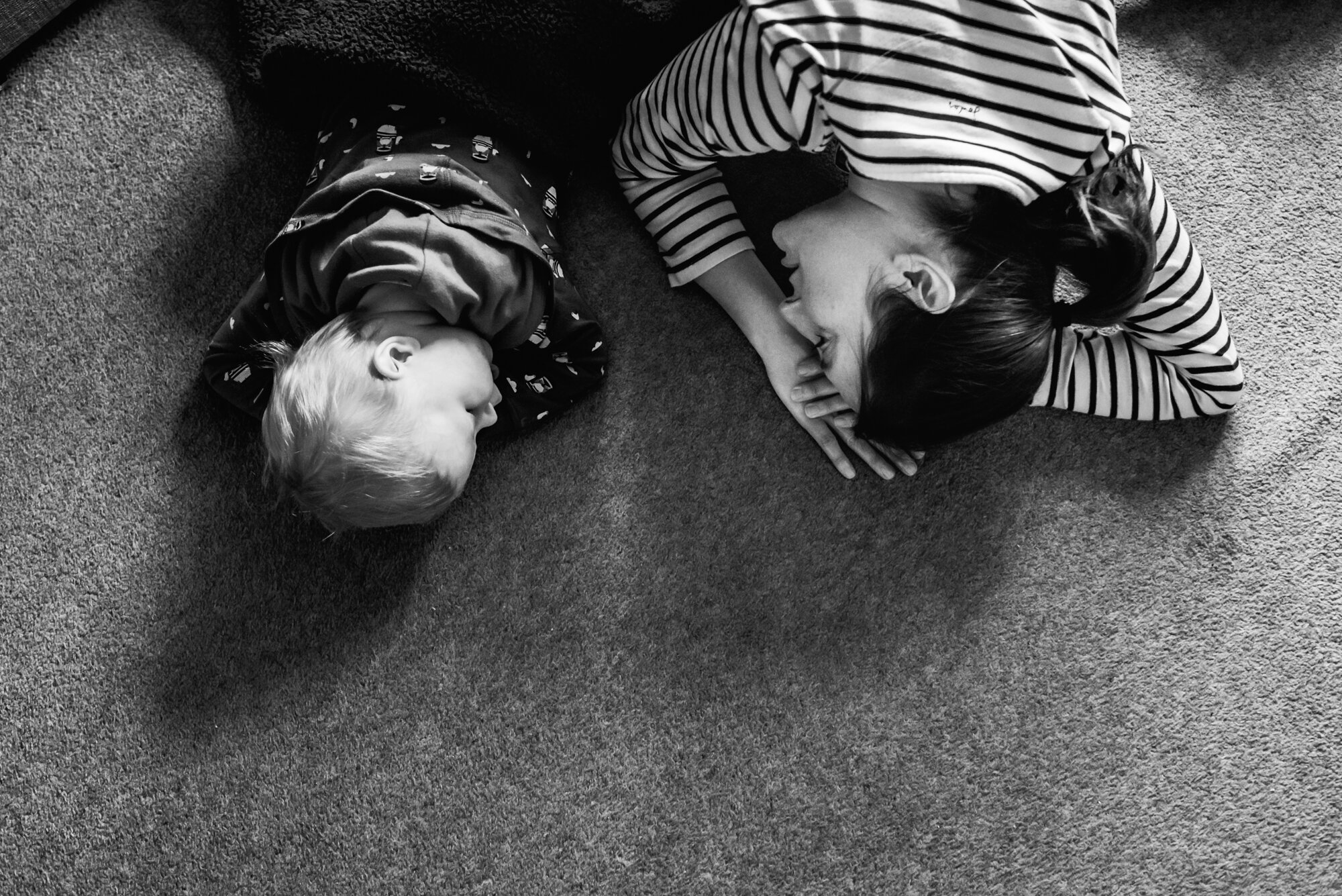  Black and white photo of a mother in a stripy top laying on the floor with her toddler son in a car cardigan. Both have their faces facing each other. 