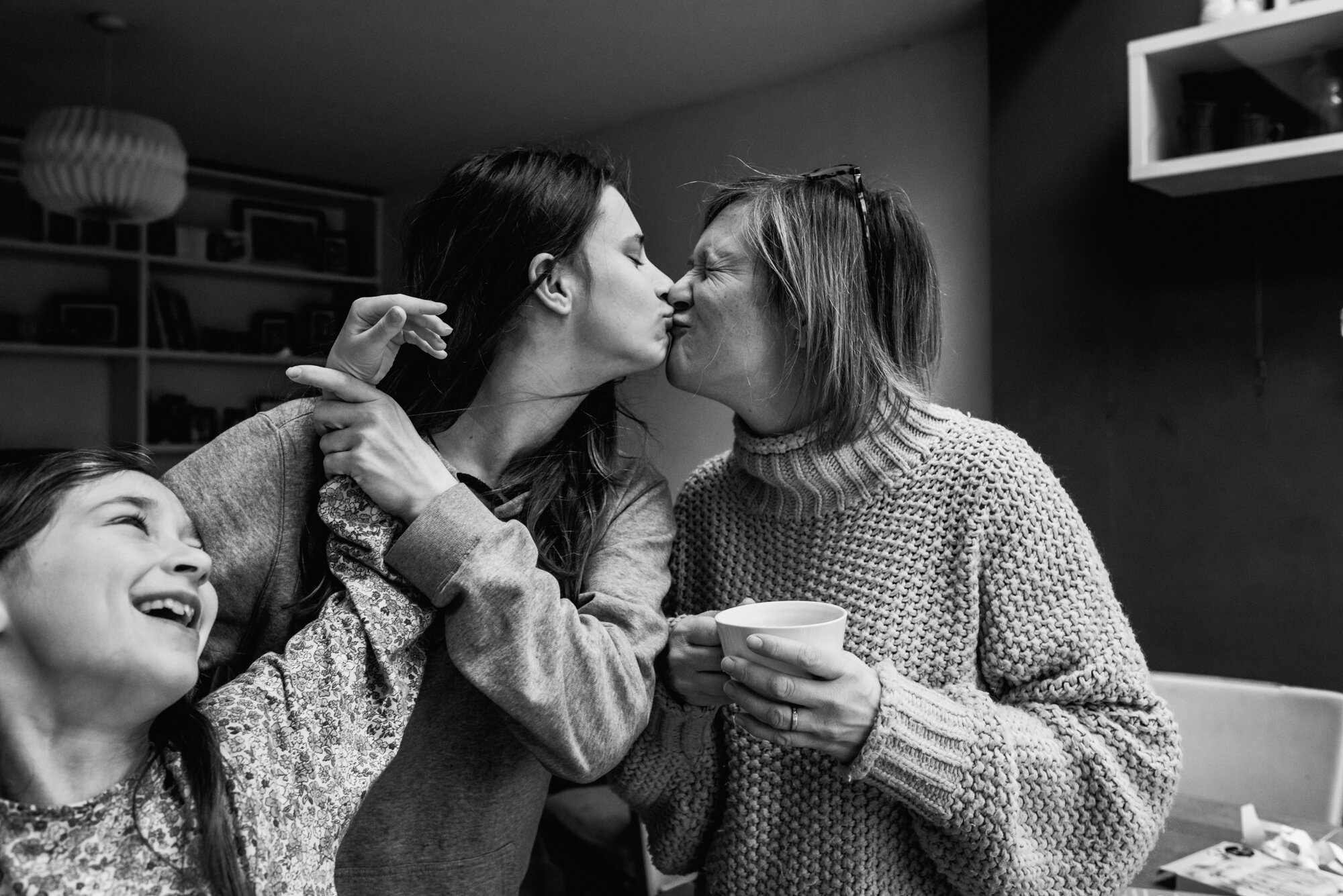  A teenage girl gives her mum a kiss as she holds a cup of tea in her hand and her sister in a floral top leans in with her arm up against her sister's hair in a day-in-the-life family photoshoot at home in St Albans, Hertfordshire 