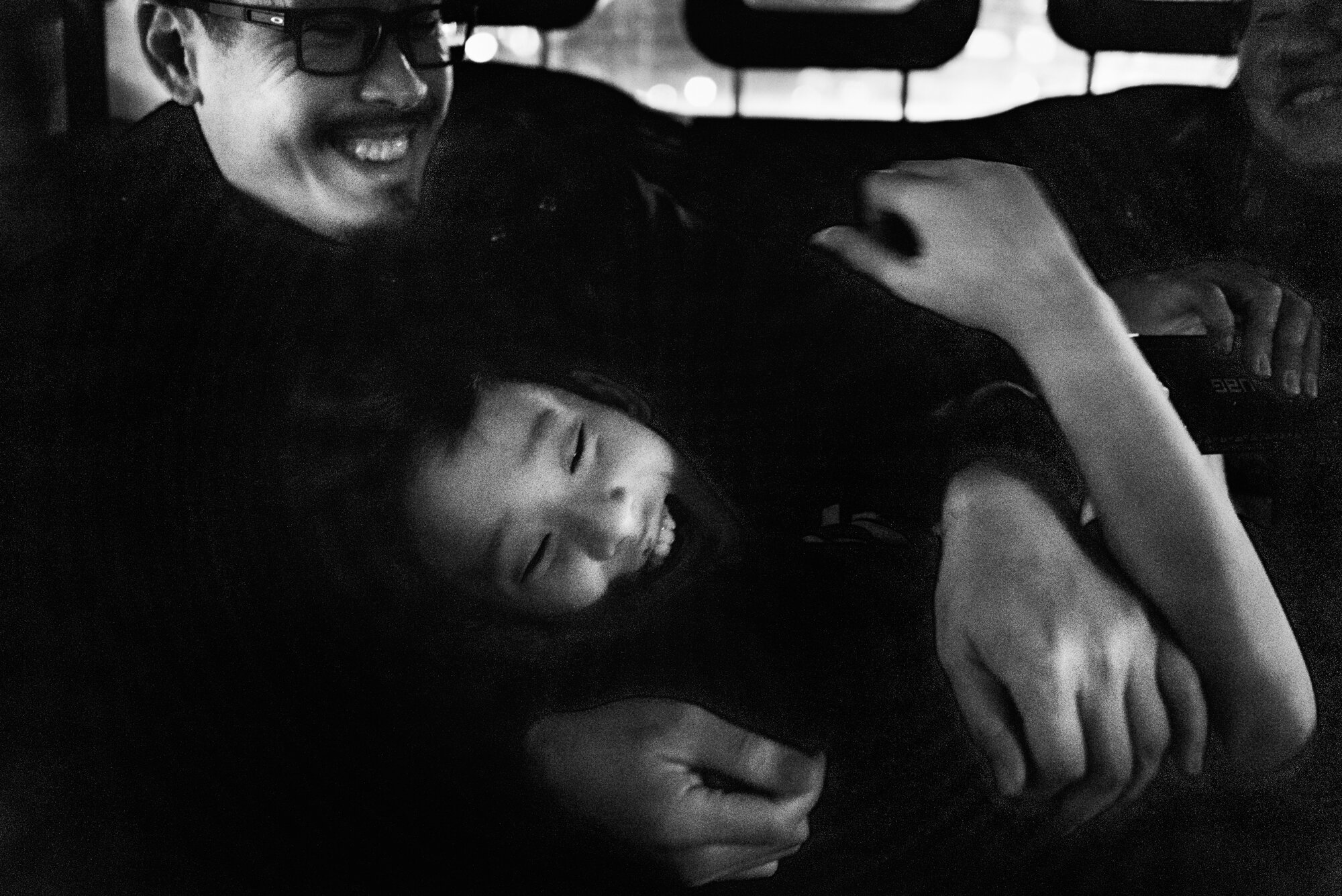  Black and white photo of a  child playfully tussle with his dad in the back of a London cab watched by his mother. 