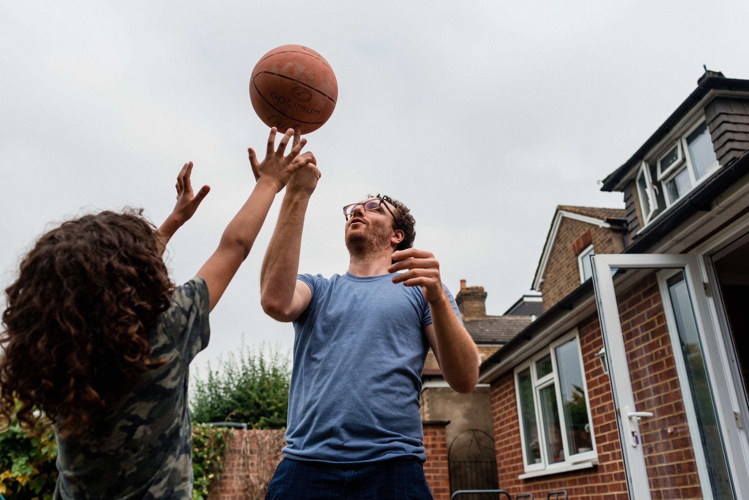  A male adult and child with dark curly hair tackle a basketball between each other's hands outside in the garden in a documentary family session in Hampton, London. 