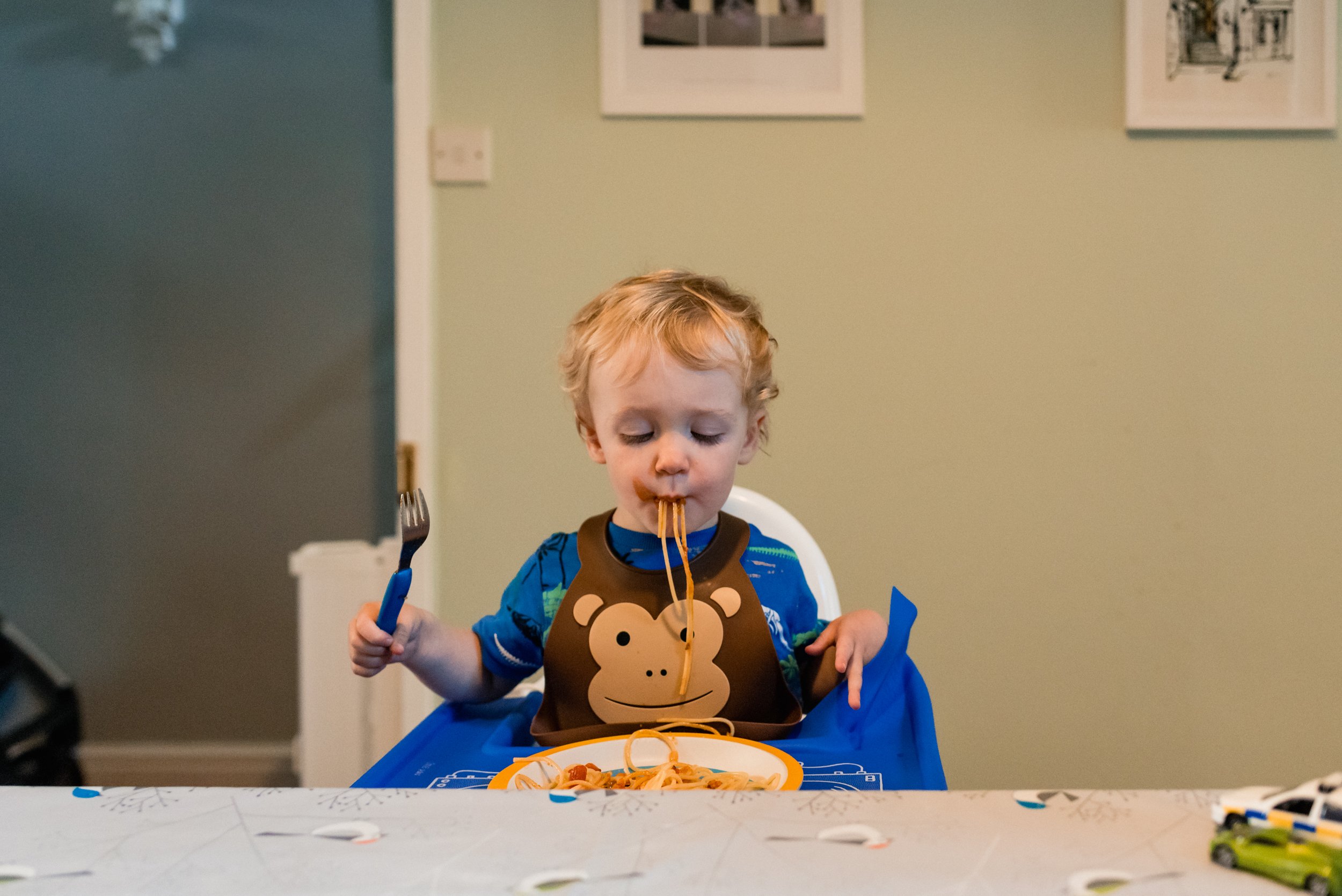  A young toddler in a monkey bib with a fork in his hand eats spaghetti by sucking the pasta through his lips. 