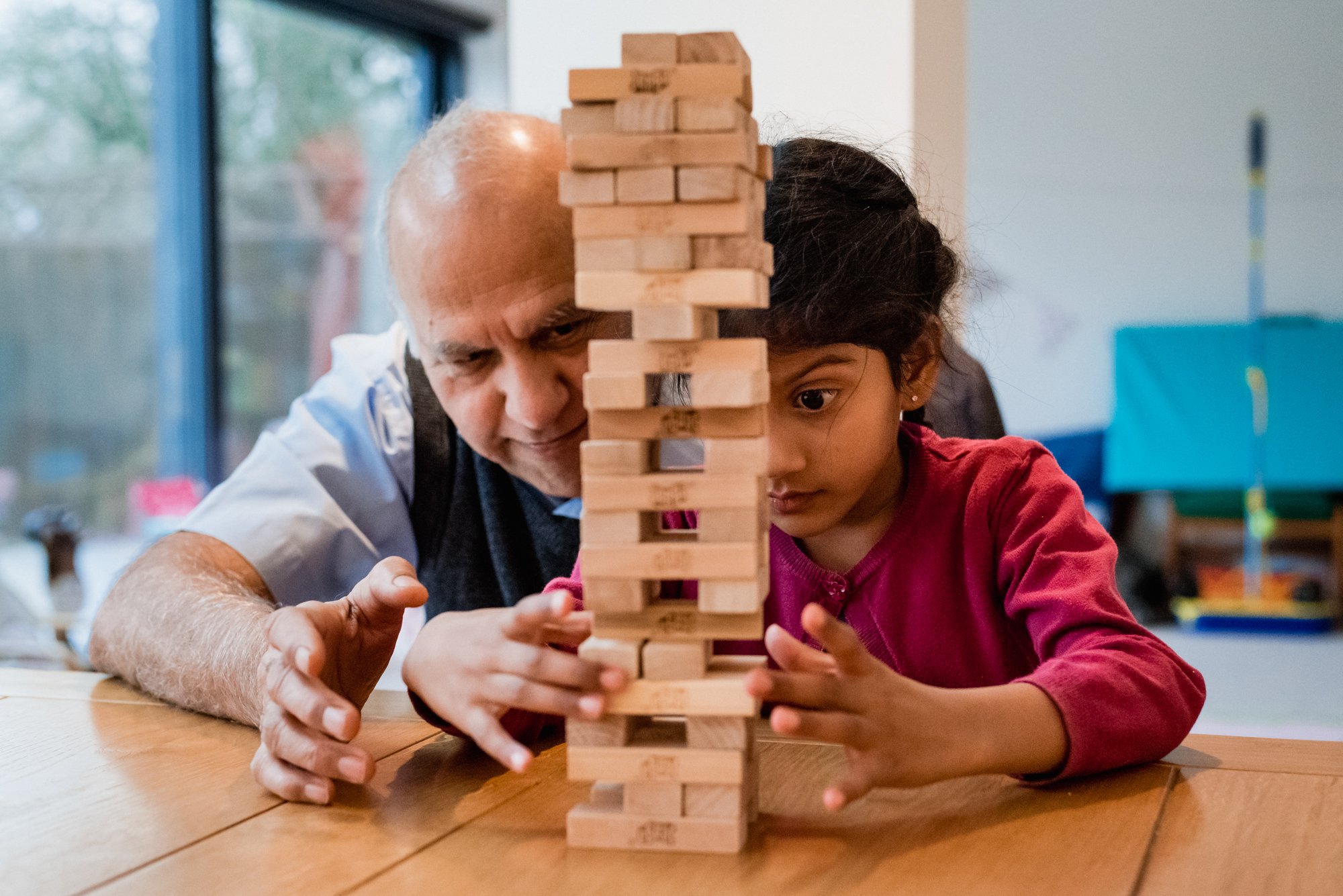 An Asian granddad and his grandchild in a pink cardigan, both of Indian origin, playing a game of jenga together. 