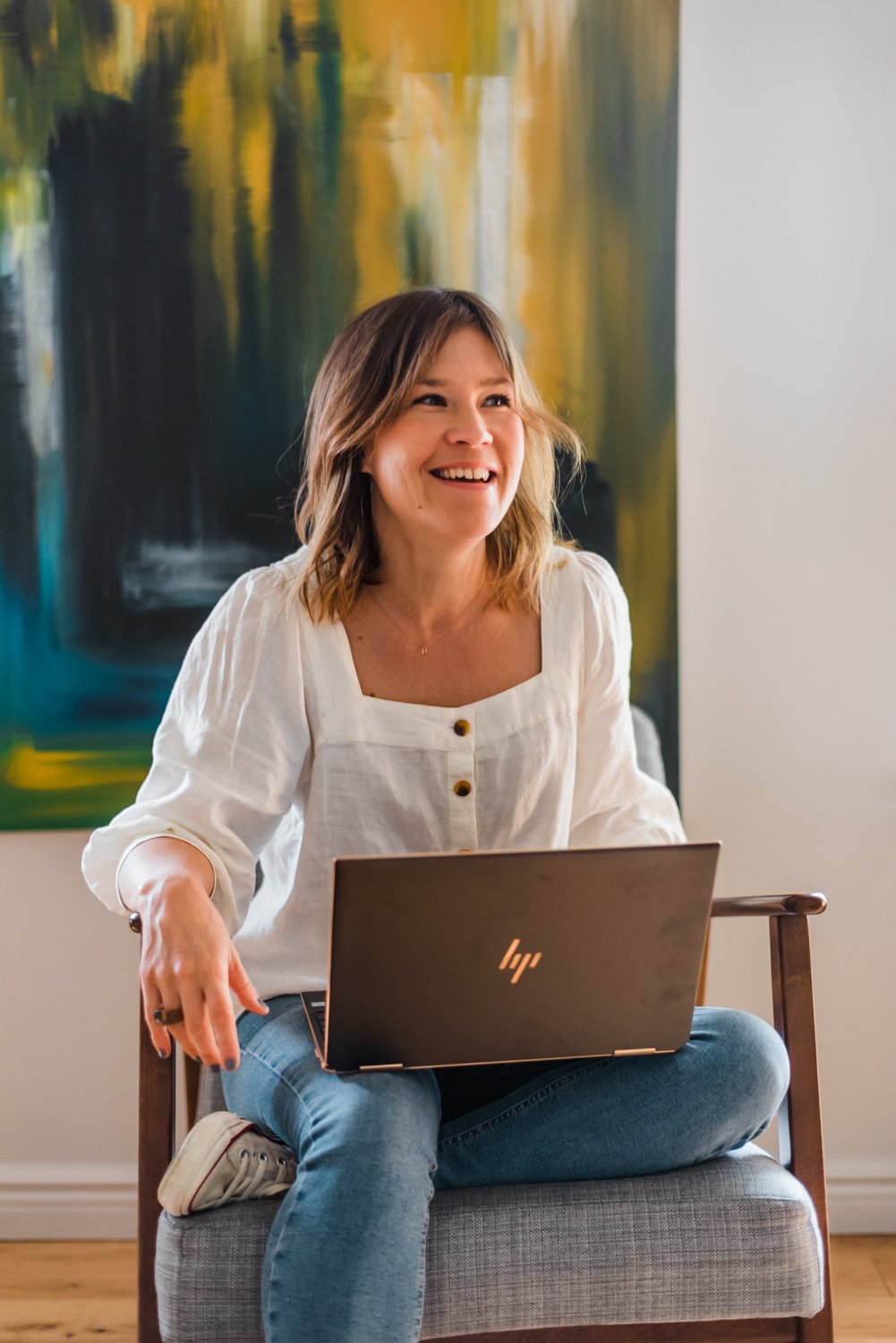  Founder of Copy Kooks, Tam Henderson, sitting in front of artwork with her laptop in a relaxed portrait. 