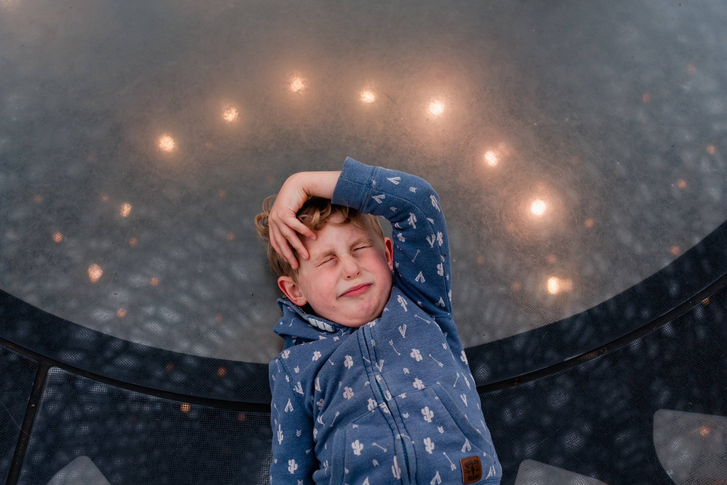  A boy in a blue cardigan decorated with white cacti is lying down on an opaque glass surface with his eyes tightly shut and one arm curved over his hear. The glass under him is reflecting the lights above and appear around his head, and the beehive 