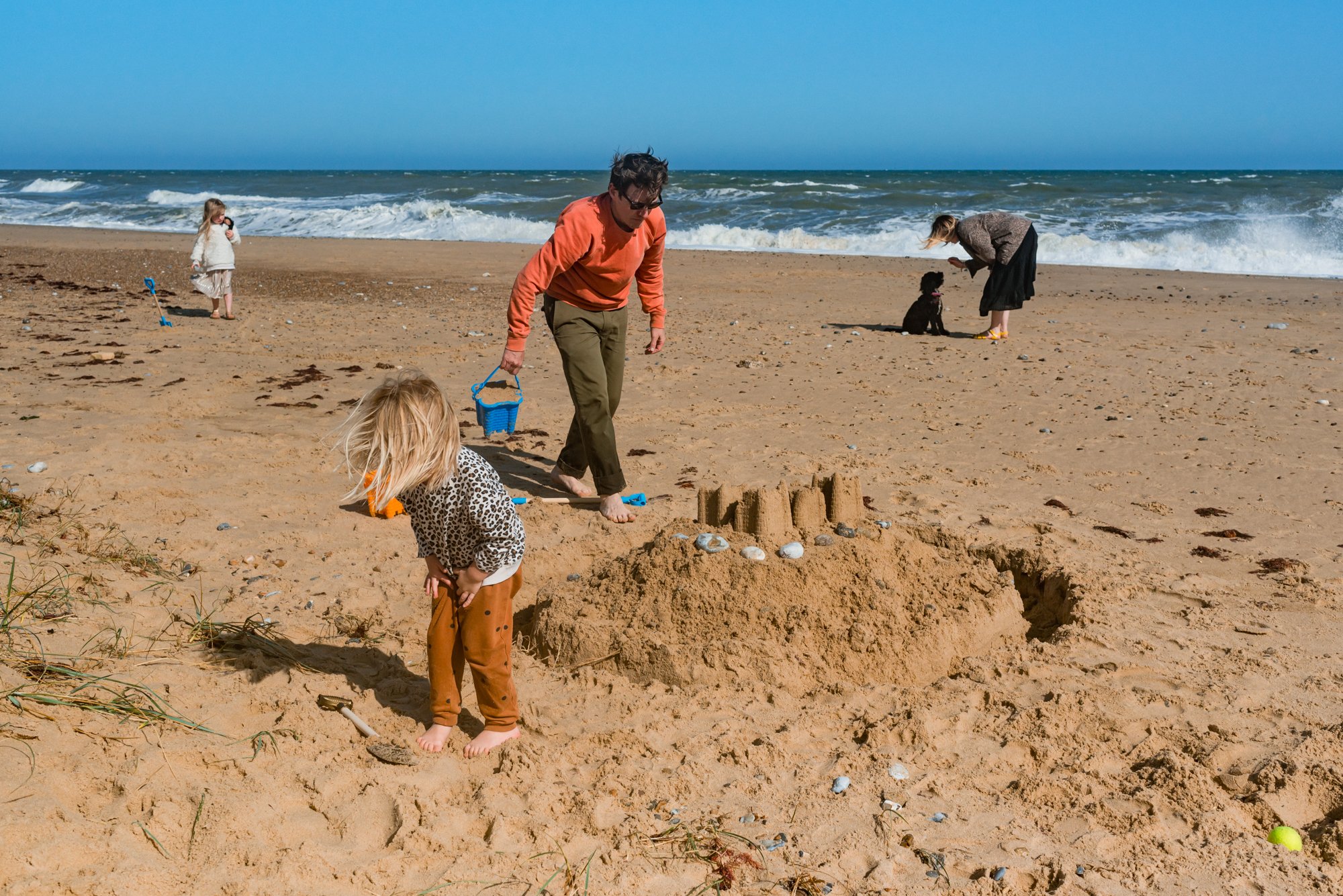  A family of five having a day out at Winterton beach, Norfolk. There is a sandcastle in the foreground and a man in orange top carrying a blue bucket walking towards the sandcastle. A woman is in the distance bending down to her black dog, and two c