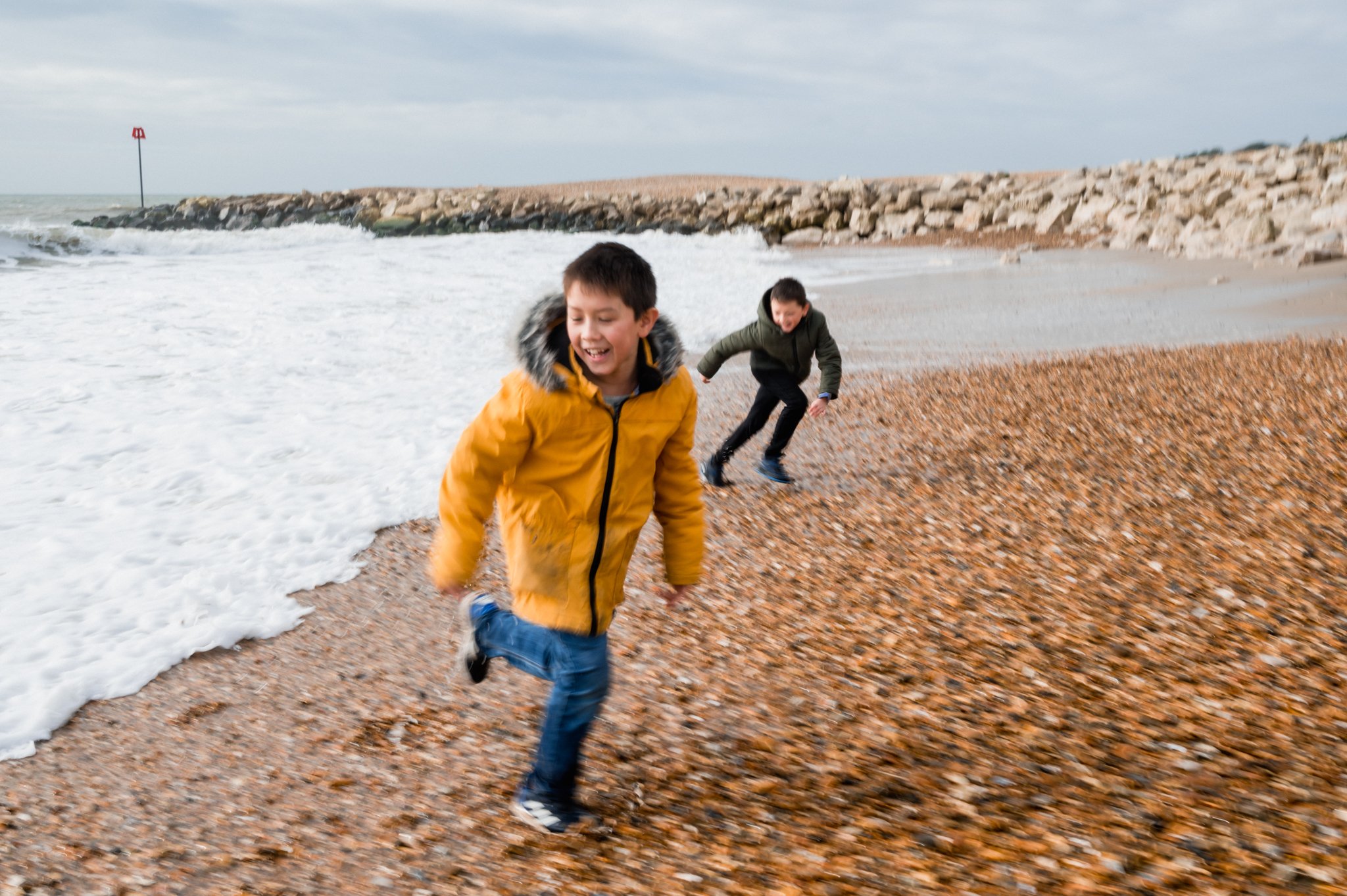  A child in a yellow winter coat and another child behind him run away from the waves at a pebble beach in Norfolk. 