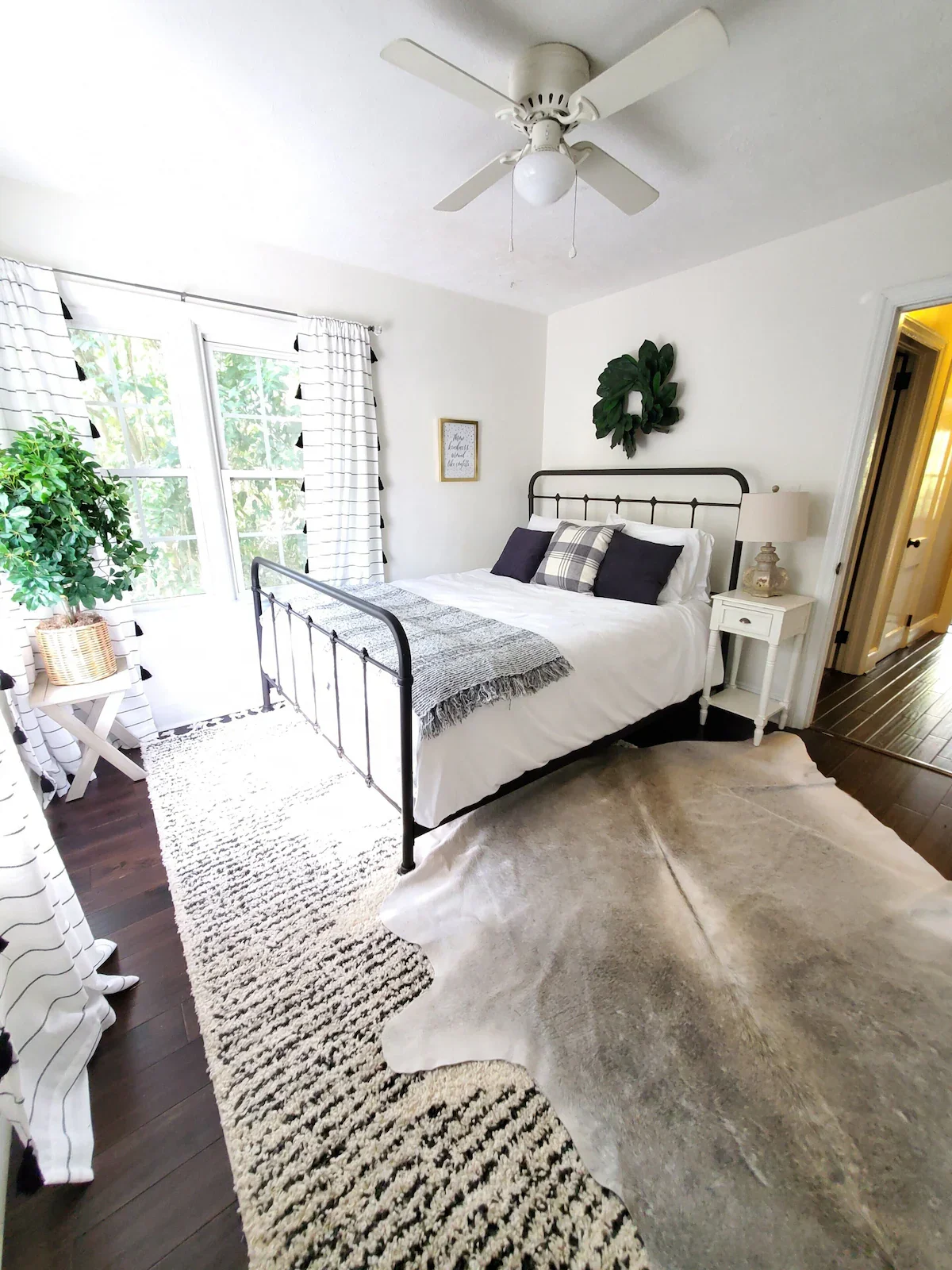 Bright bedroom with white walls, black metal bed frame, white bedding, plaid and solid pillows, white nightstand with lamp, large window with striped curtains, potted plant, wall decor, ceiling fan, wooden floor with a rug and animal hide.