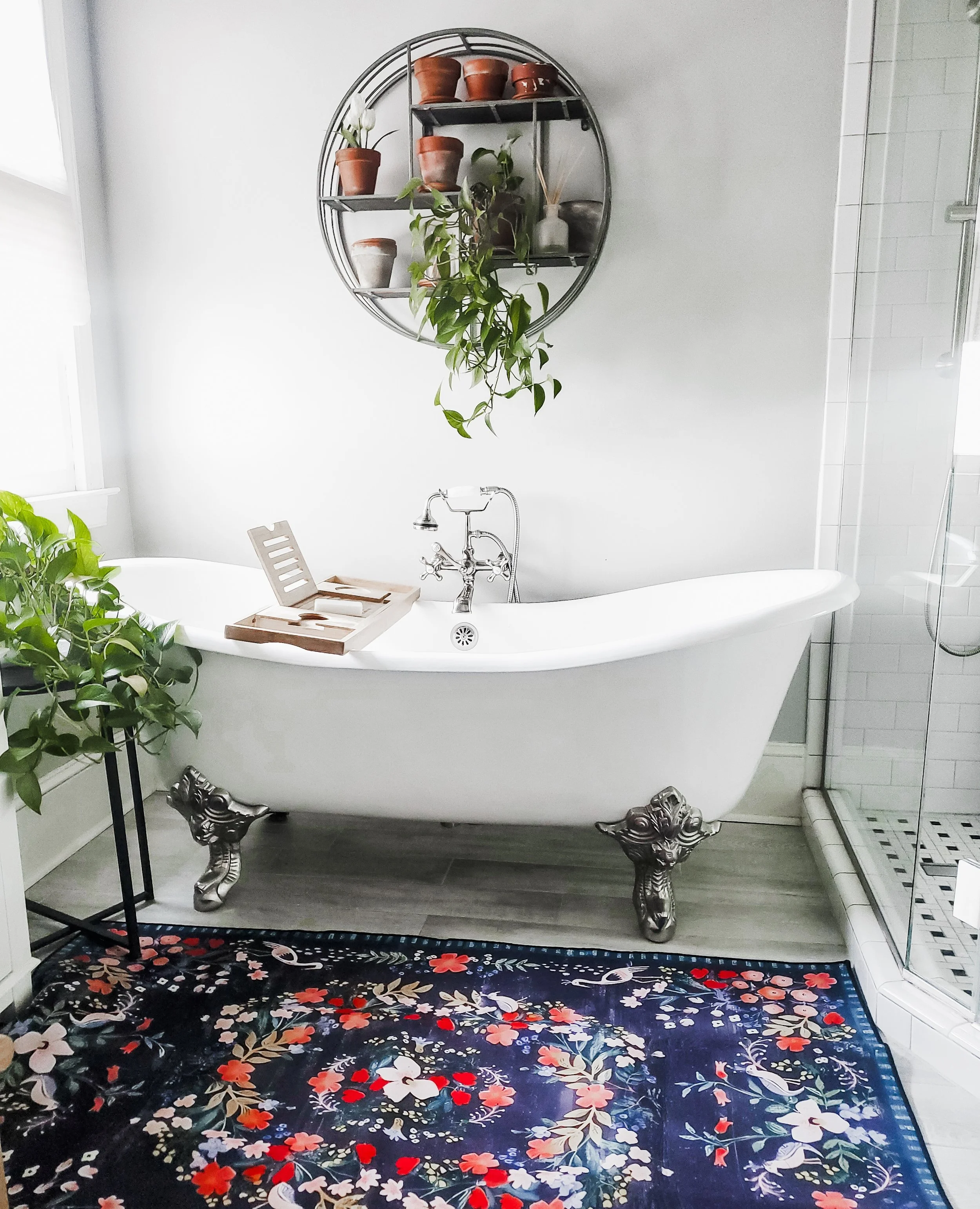 Vintage white clawfoot bathtub with curved metal clawfoot legs in a modern bathroom, with a dark floral rug, potted plants, a wall shelf with pots and decor, and a glass shower enclosure.