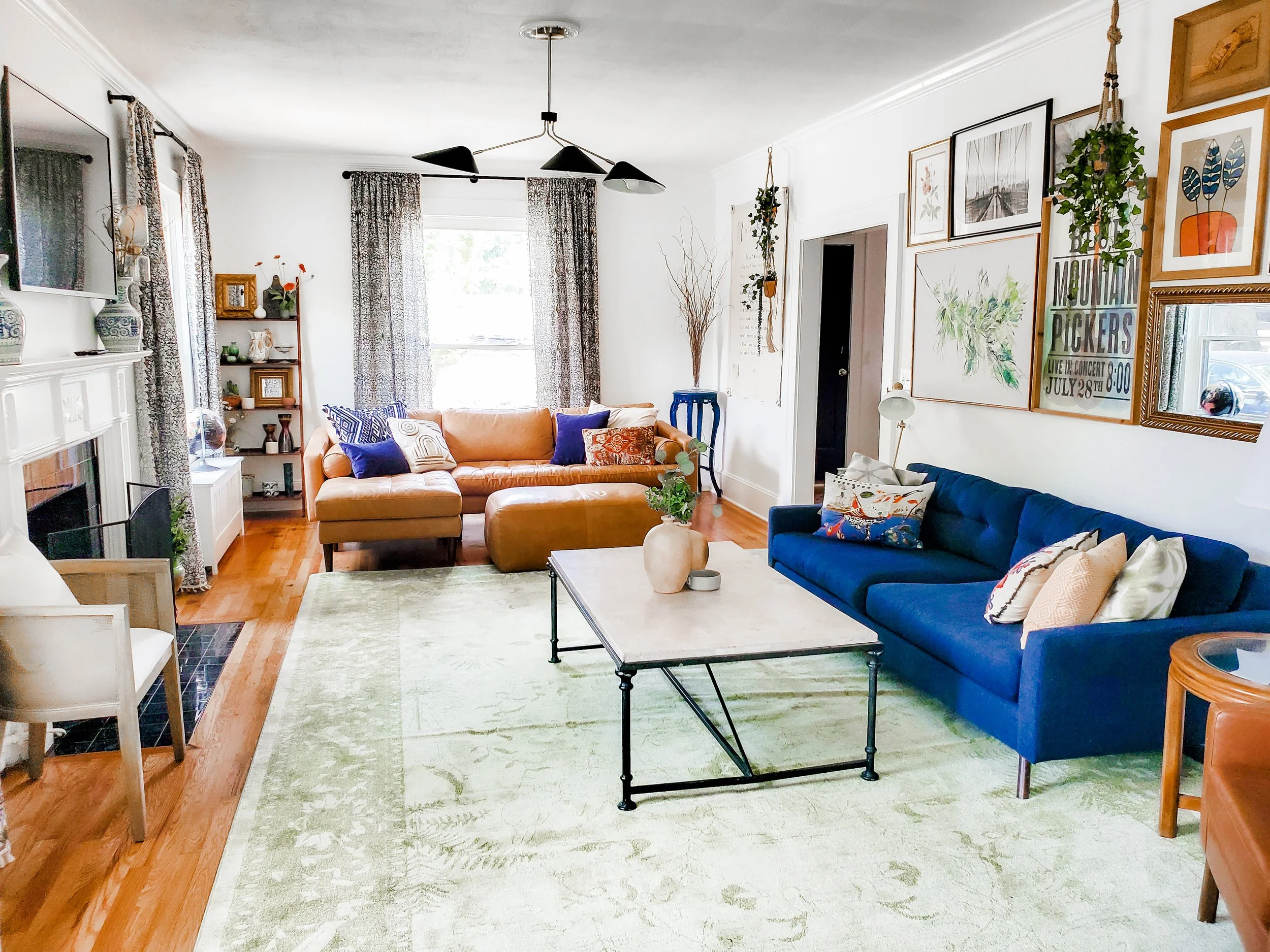 Living room with two sofas, one tan and one blue, a white coffee table, a green rug, and various decorative pillows. There is a fireplace on the left with vases and art on the mantel, and a bookshelf with decor items in the back corner. The walls are decorated with framed artwork, plants, and photographs. A window with patterned curtains lets in natural light.