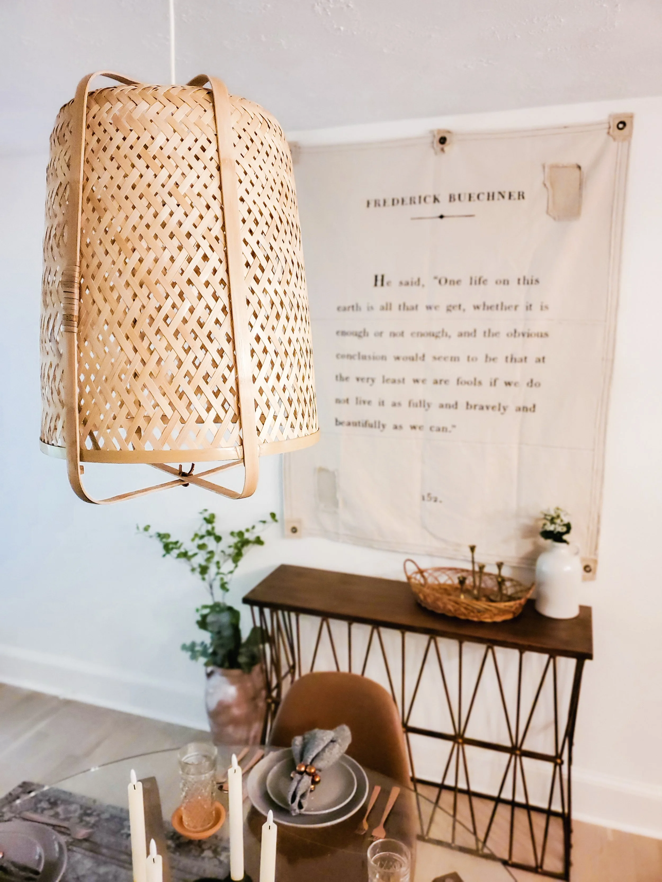 Interior dining room with hanging wicker light fixture, a table set with candles, plates, and glasses, a wooden sideboard, a decorative basket, a small plant, and a wall hanging with text.