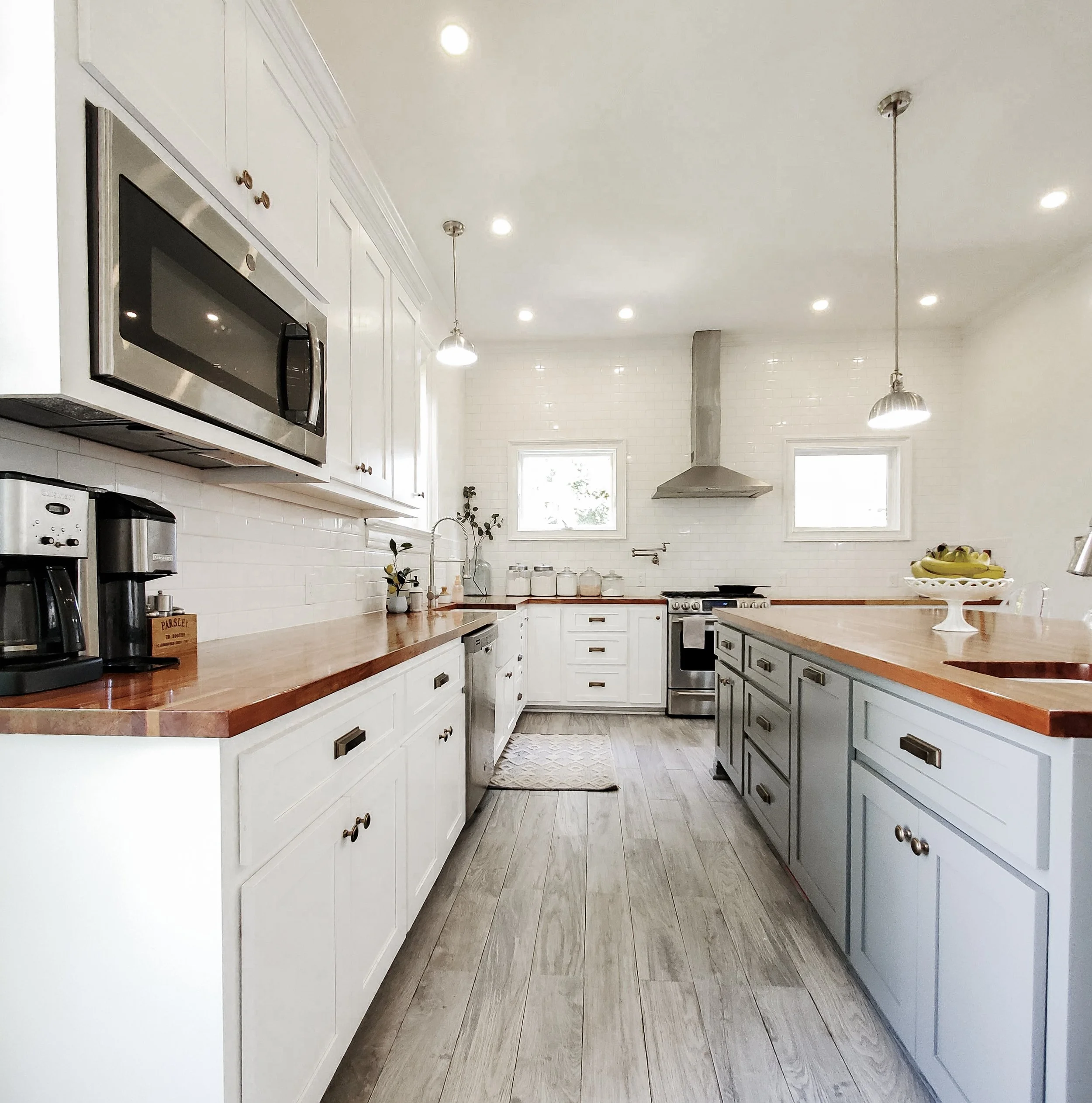 Bright kitchen with white cabinets, wood countertops, stainless steel appliances, two small windows, pendant lights, and a wooden floor.