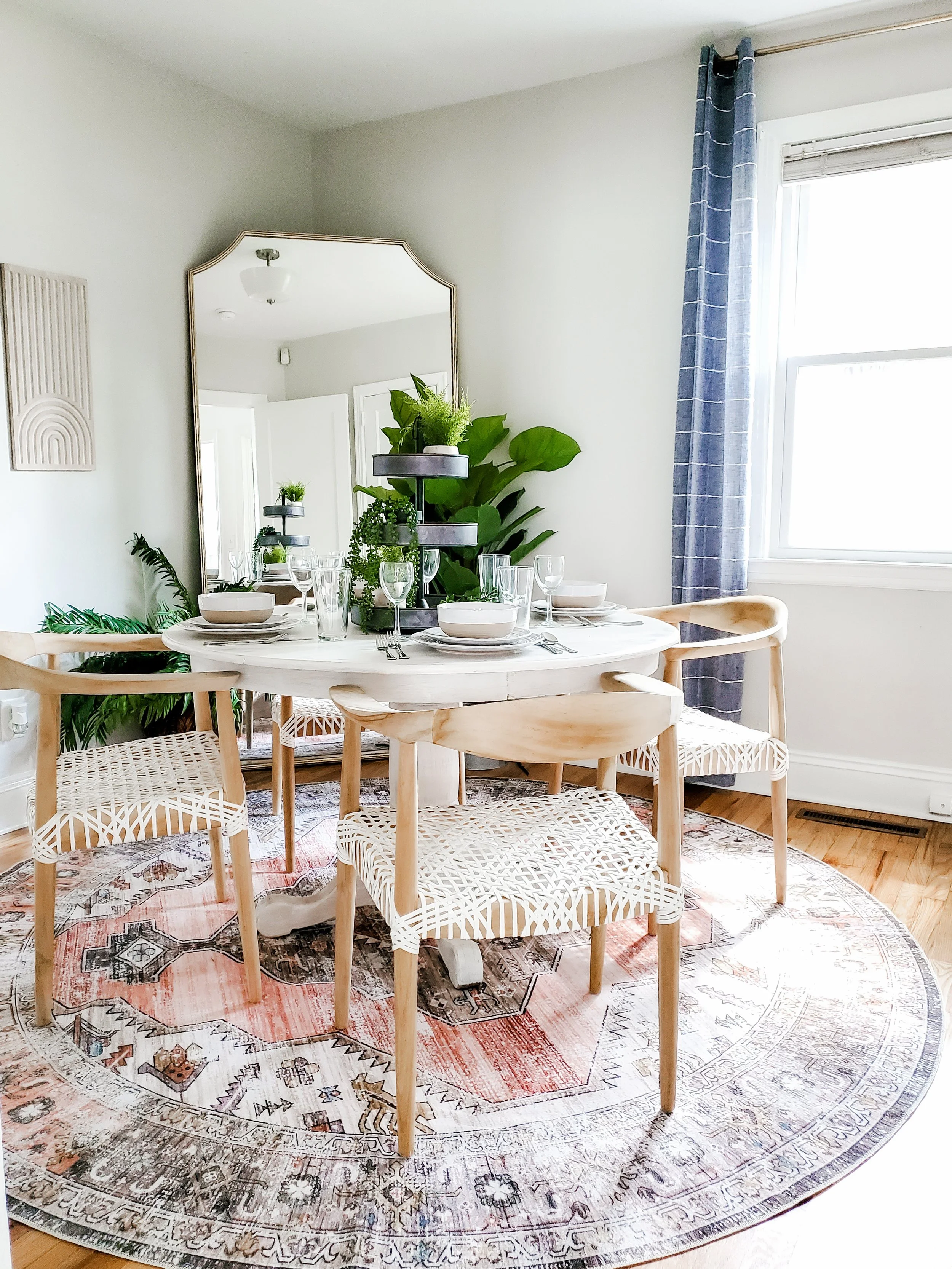 A dining room with a round white table, four wooden chairs with woven seats, a mirror, and greenery. The table is set with bowls, glasses, and silverware, and the room has a colorful rug, blue curtains, and a window with blinds.