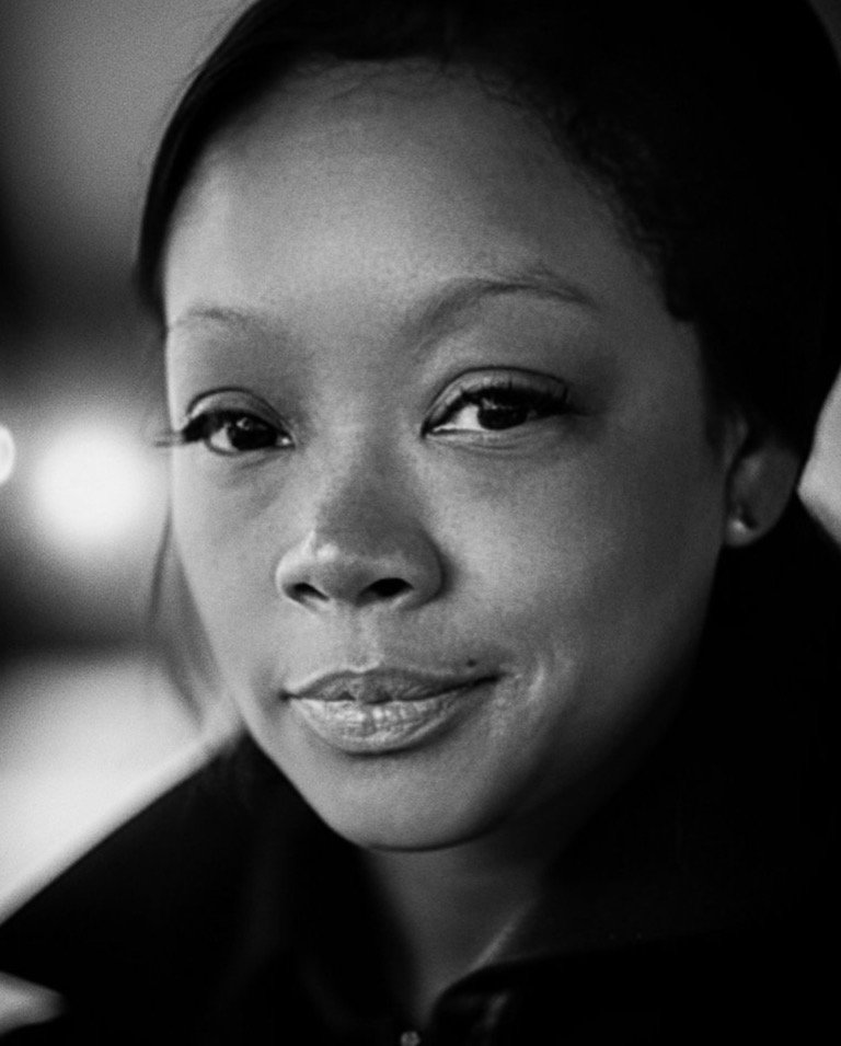A close-up black and white photograph of a young African American woman with smooth skin, full lips, and almond-shaped eyes, looking directly at the camera with a slight smile.