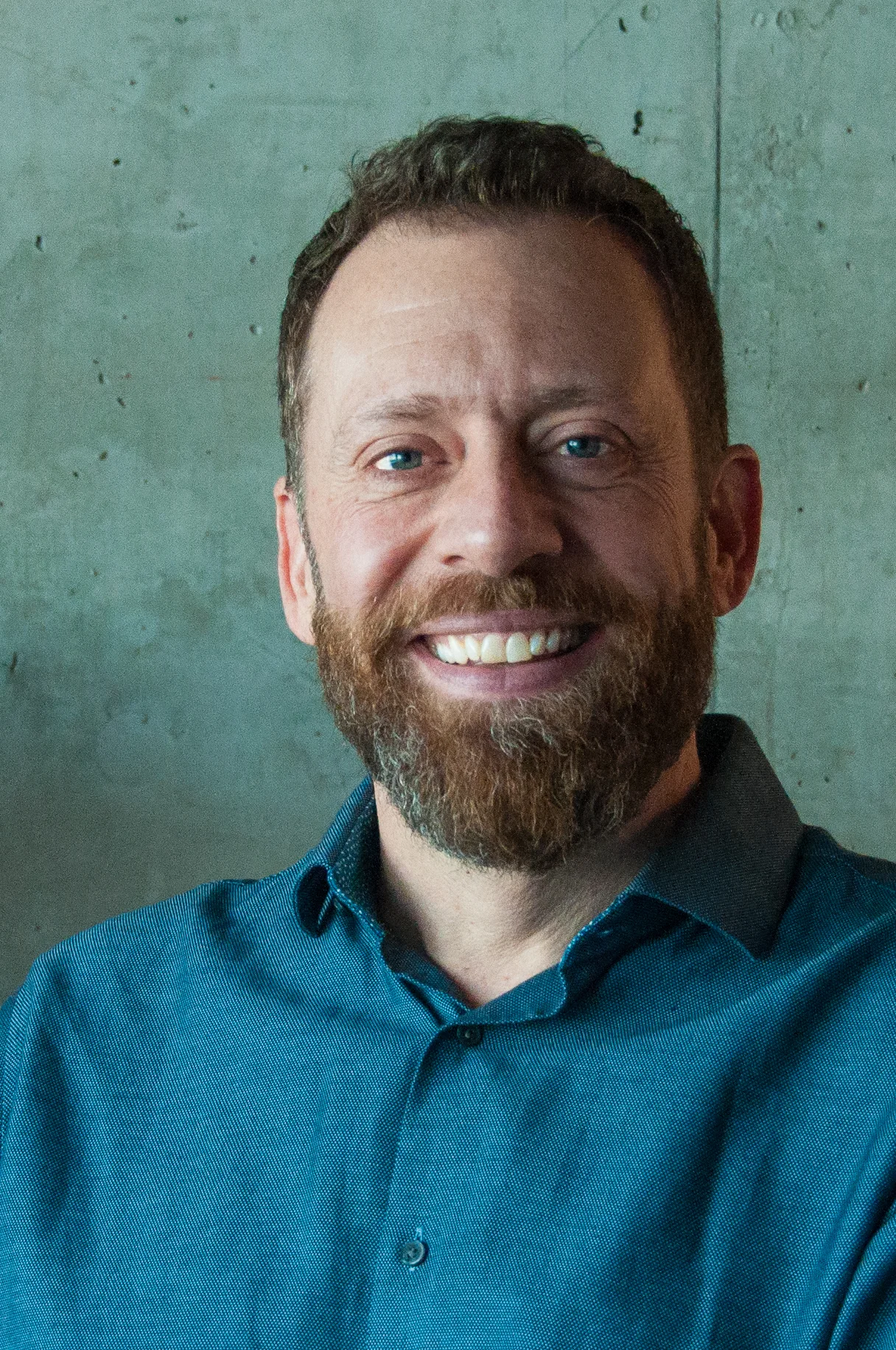 A man with short brown hair and a beard, smiling, wearing a blue collared shirt, standing against a plain gray/green wall.