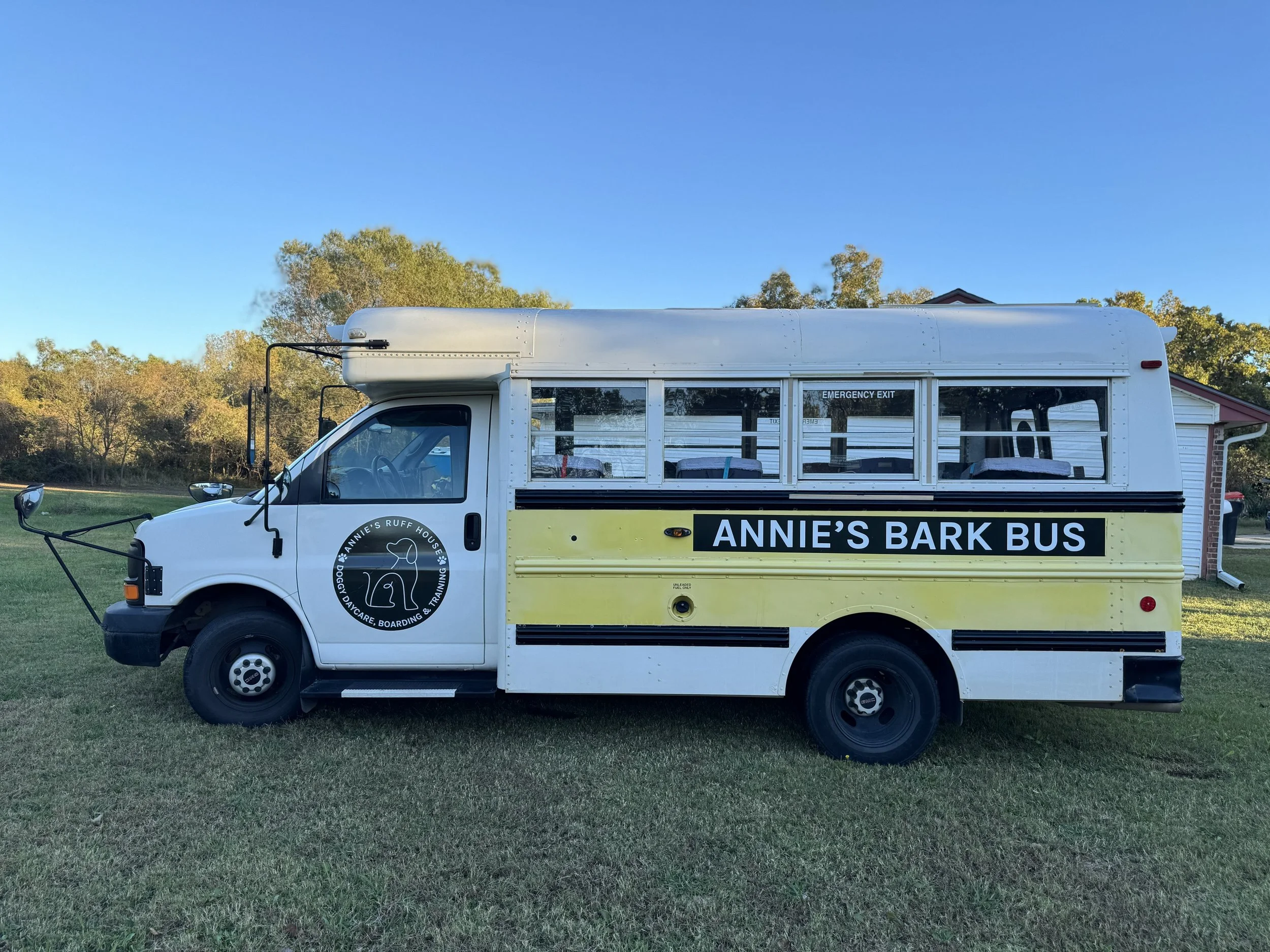 Custom bus with signage for Annie's Bark Bus, a dog daycare and boarding service, parked on grass with trees and a house in the background.