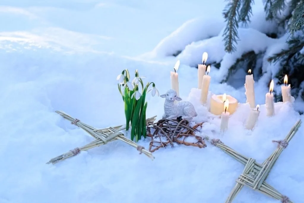 A statue of a lamb sits on top of a woven star beside blooming snowdrops and candles burning candles on a snowy background