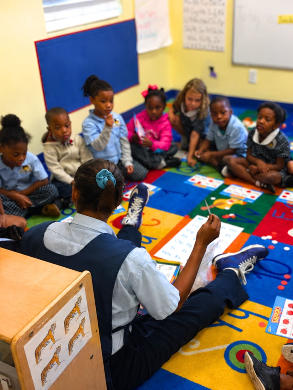 Teacher guiding preschoolers through an alphabet and spelling activity on a classroom rug.