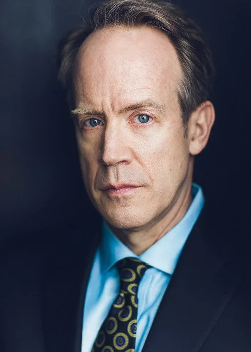 A middle-aged man with blue eyes and light brown hair, dressed in a dark suit, light blue shirt, and patterned tie, looking serious against a dark background.