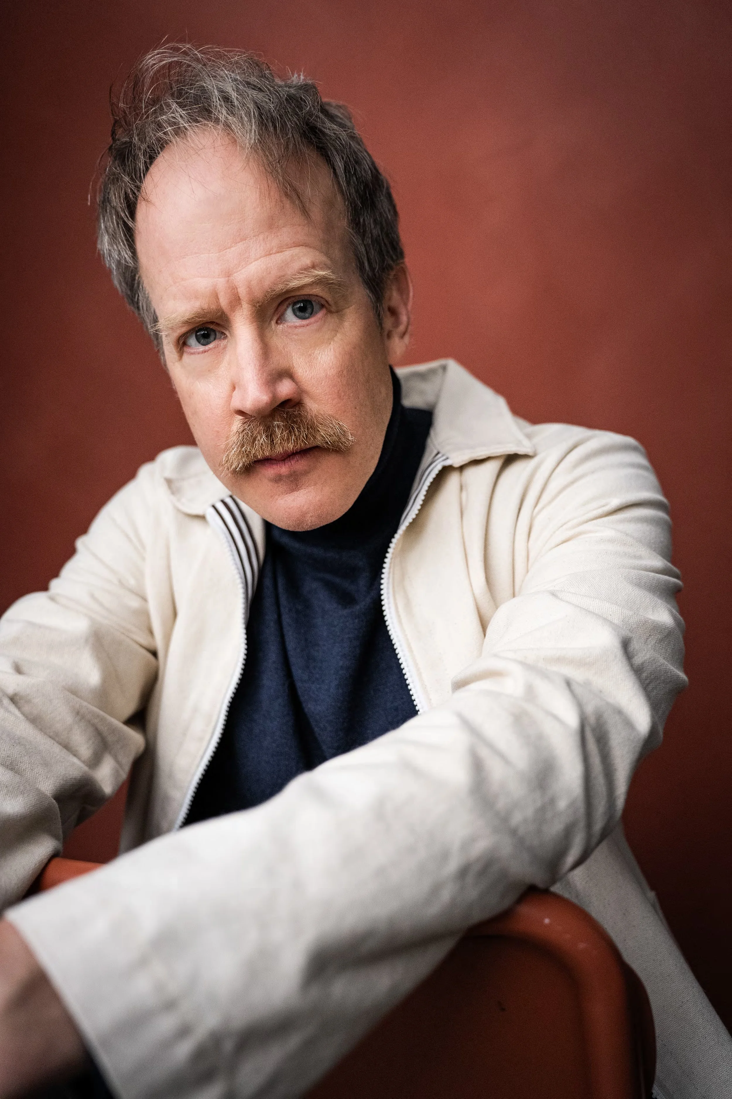 Close-up portrait of a man with blue eyes, a mustache, and messy gray hair, wearing a beige jacket over a dark shirt, against a reddish-brown background.