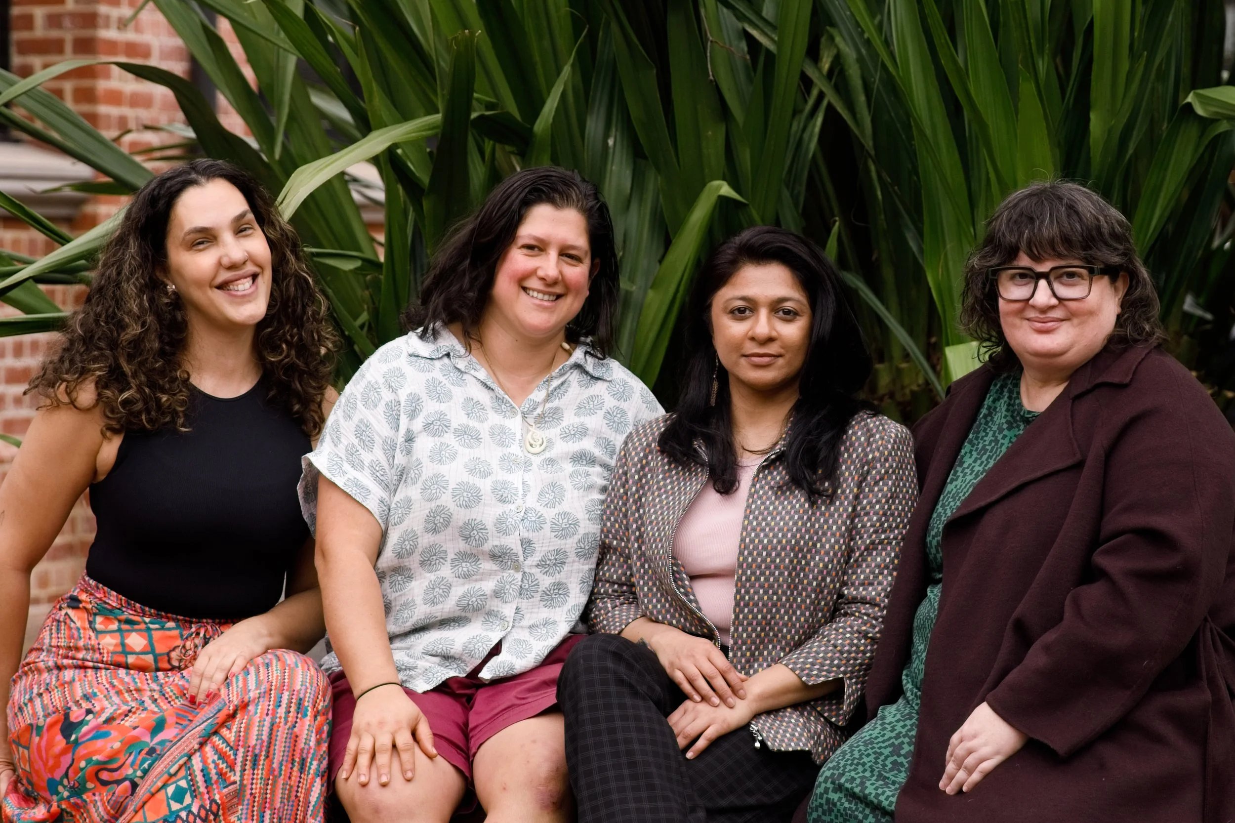 Four cis women sit together outdoors in front of lush green plants, smiling at the camera. They're dressed in colourful, patterned clothing and dark outer layers.
