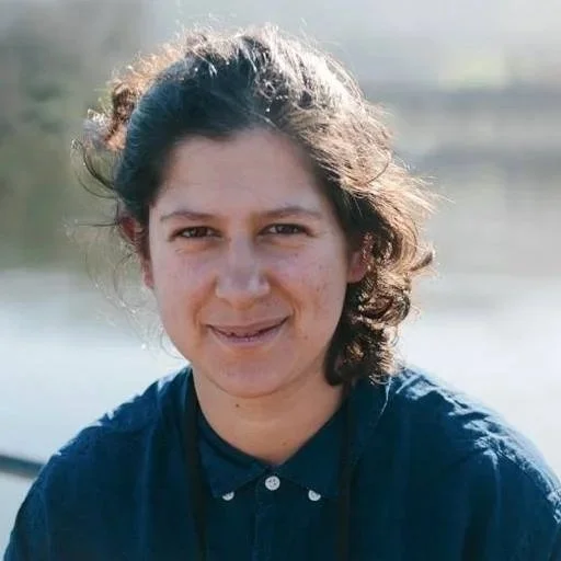 Elena smiling at the camera with dark, windswept hair wearing a dark collared shirt outdoors.