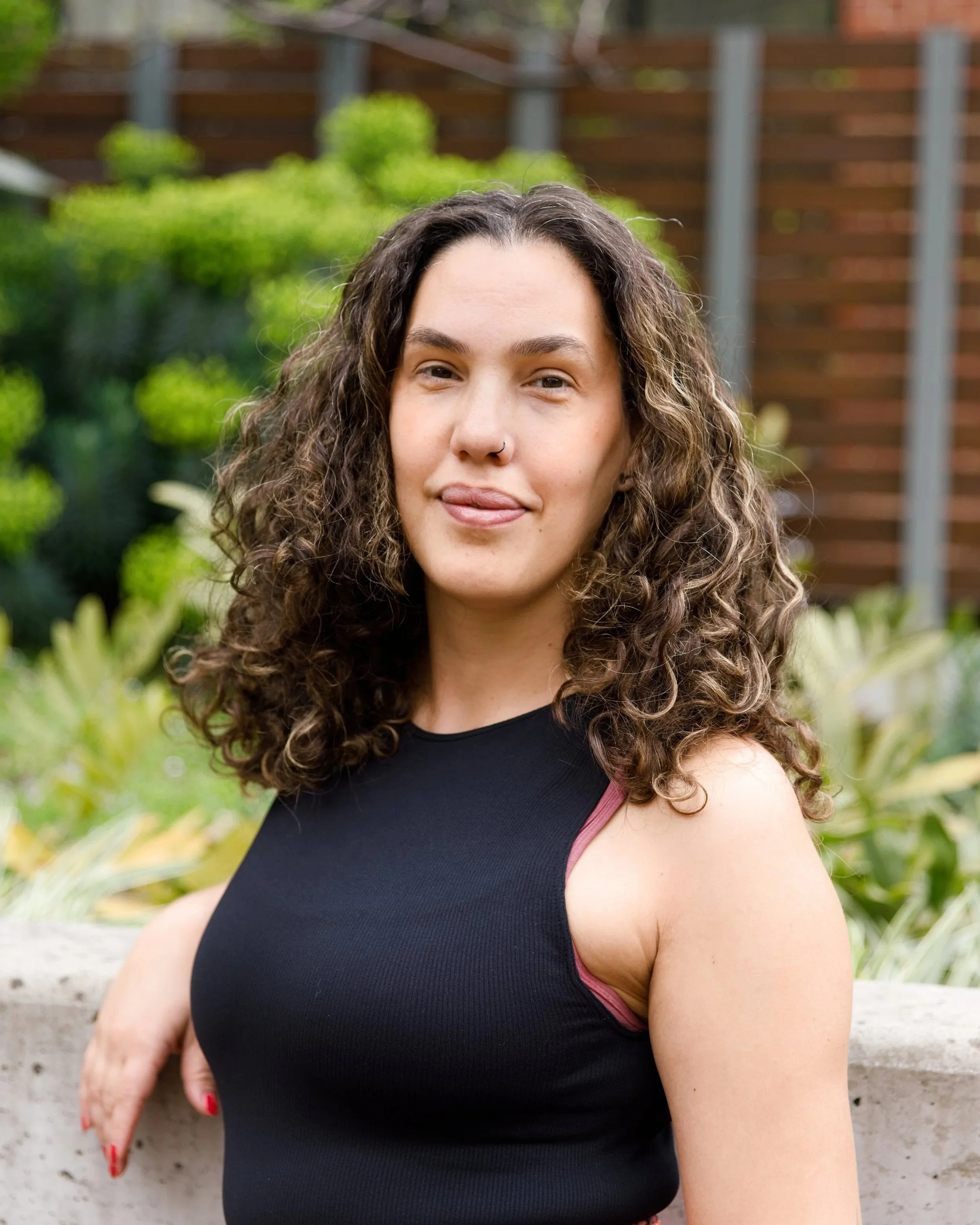 A woman with shoulder-length, curly brown hair and a nose ring smiles faintly as she leans against a concrete wall in a garden. She wears a sleeveless black top and colourful patterned trousers.