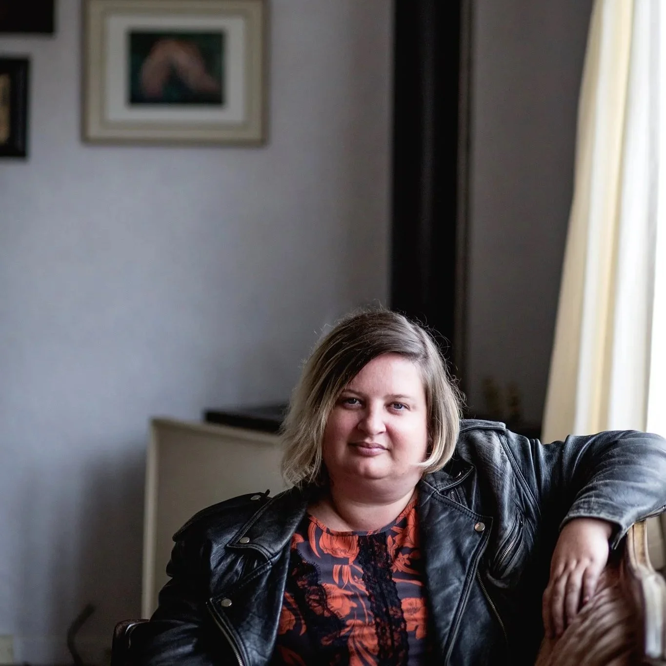 Megan with short, light brown hair sits on a couch indoors, wearing a black leather jacket over a patterned top.