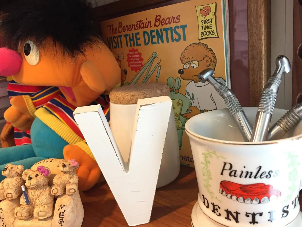 Sesame street's Ernie laying against Berenstein Bears "Visit to the Dentist" children's book, next to a "Painless Dentist" teacup holding three drills inside it, one etched with Dr. Viera's father, Claudio Rivera's name.