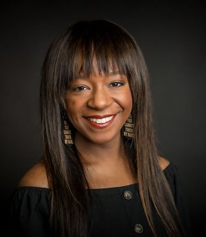 An image description of the photo you sent (textual description of what we see in the photo, for patrons who are blind or have low vision.: A woman smiles at the camera against a black background. She wears a black blouse and black earrings that say No. You cannot touch my hair.