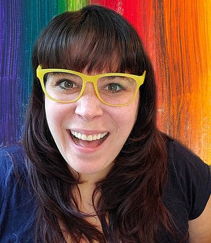 Katherine (she|they), a white person with burgundy hair worn down with bangs, is wearing yellow glasses, a dark blue shirt, and smiling in front of a rainbow background. 