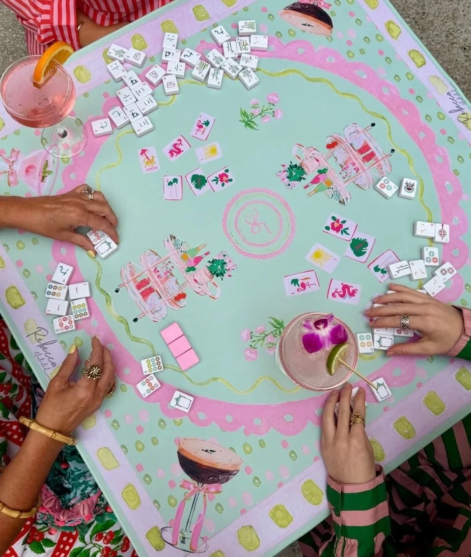 Colorful overhead mahjong table with tiles, players’ hands, and cocktails.