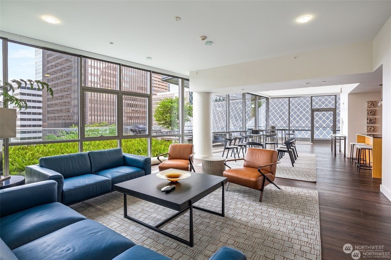 Modern lounge area with blue sofas and brown leather chairs, large windows with city view, and a dining area with tables and chairs.