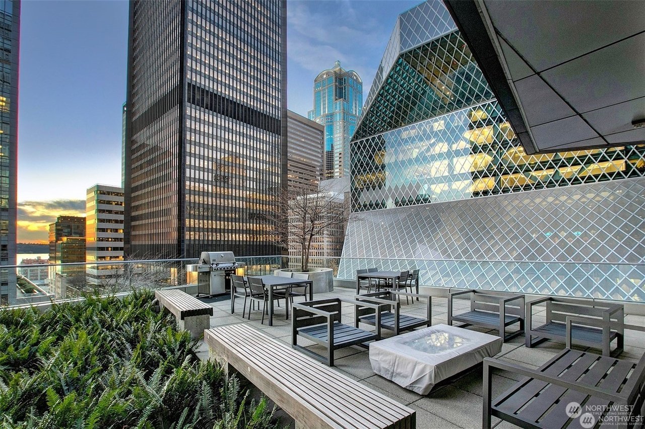 Rooftop terrace in urban setting with modern furniture, barbecue grill, and surrounding skyscrapers. View includes greenery and glass facade building reflecting evening sky.