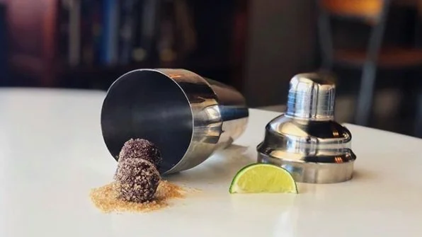 Two Mojito truffles coated in demerrara sugar outside of a cocktail shaker turned onto its side with a slice of lime nearby