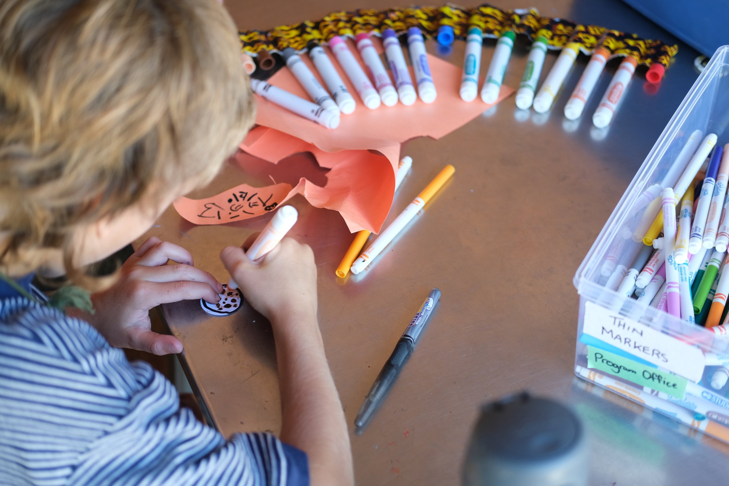 An over-the-shoulder picture of a child at a summer camp drawing on a piece of paper. It's a blond child in a blue striped shirt at an arts and crafts table, with a number of Sharpie markers arrayed in a fan in front of the child.