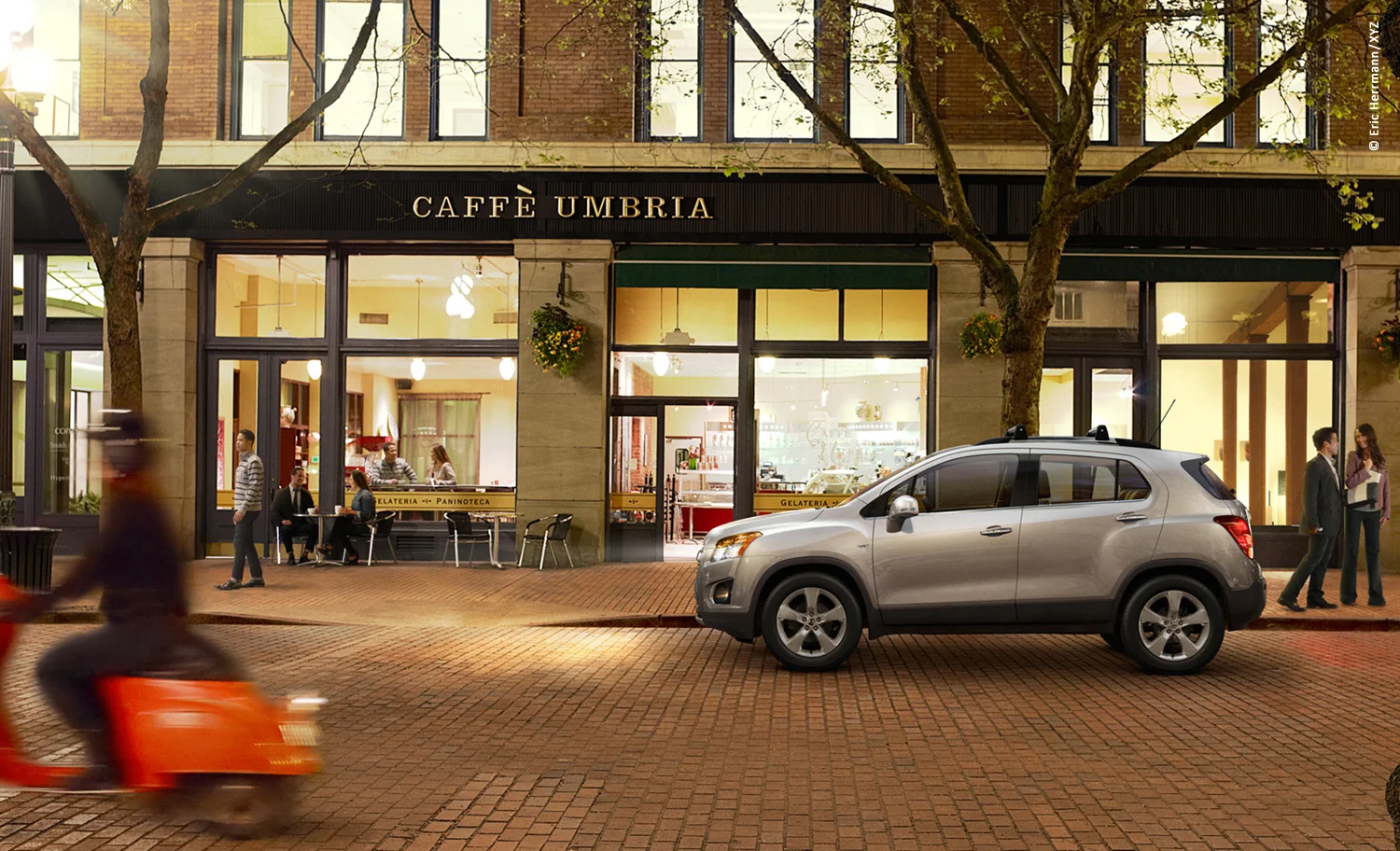 photo of night time street scene with several groups of people, a moving Vespa scooter and Chevy Trax car in front of a Italian cafe