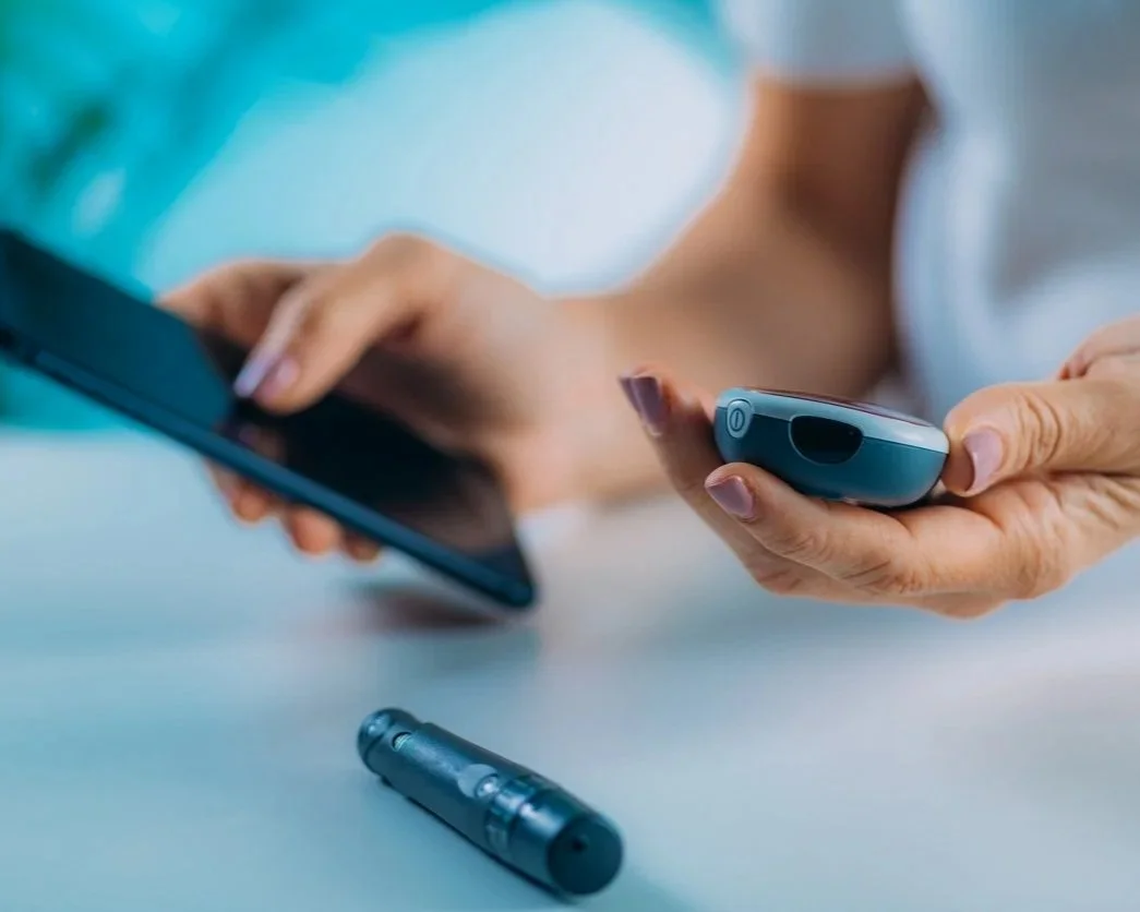 Person holding a blood glucose meter and a smartphone, with a blood glucose test strip container on the table.