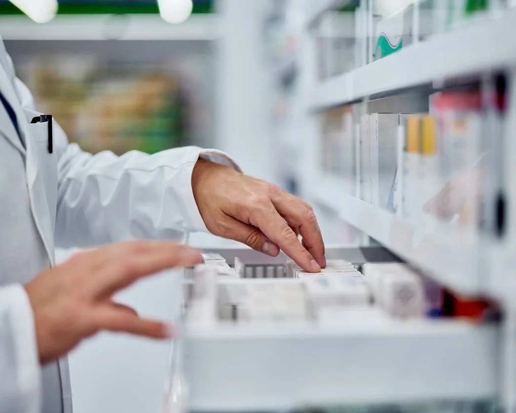 A person in a white lab coat selecting medication from a pharmacy shelf.