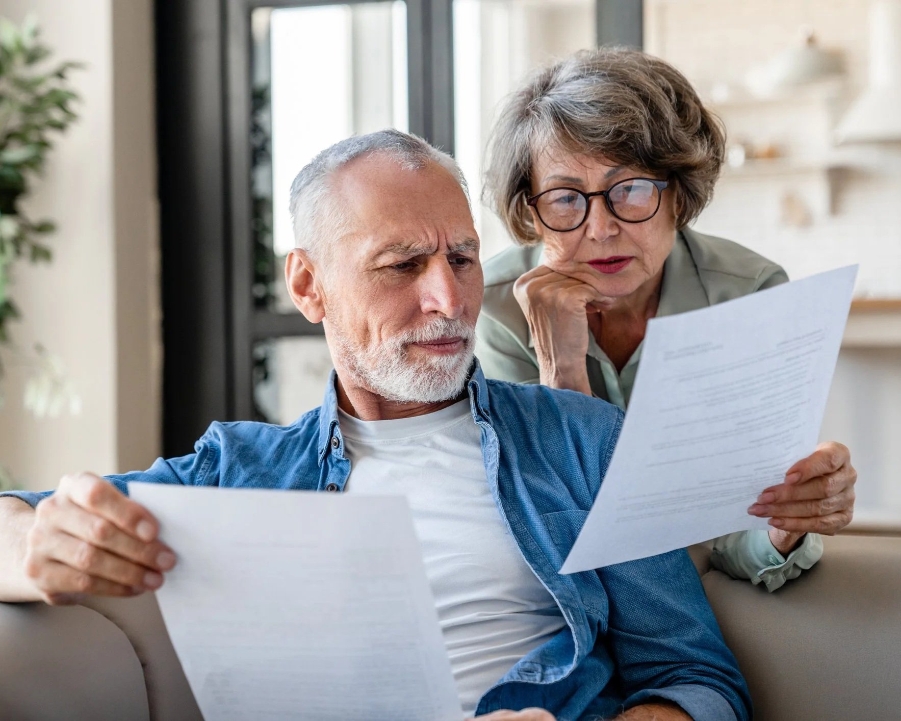 An elderly man and woman sitting on a couch, reviewing documents together in a bright room.