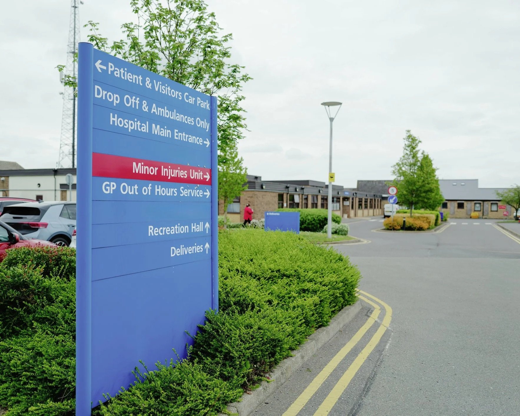 Hospital direction sign with various locations and services listed, including patient and visitor parking, ambulance drop off, main entrance, minor injuries unit, GP out of hours service, recreation hall, and deliveries, with a hospital building in the background and parked cars nearby.