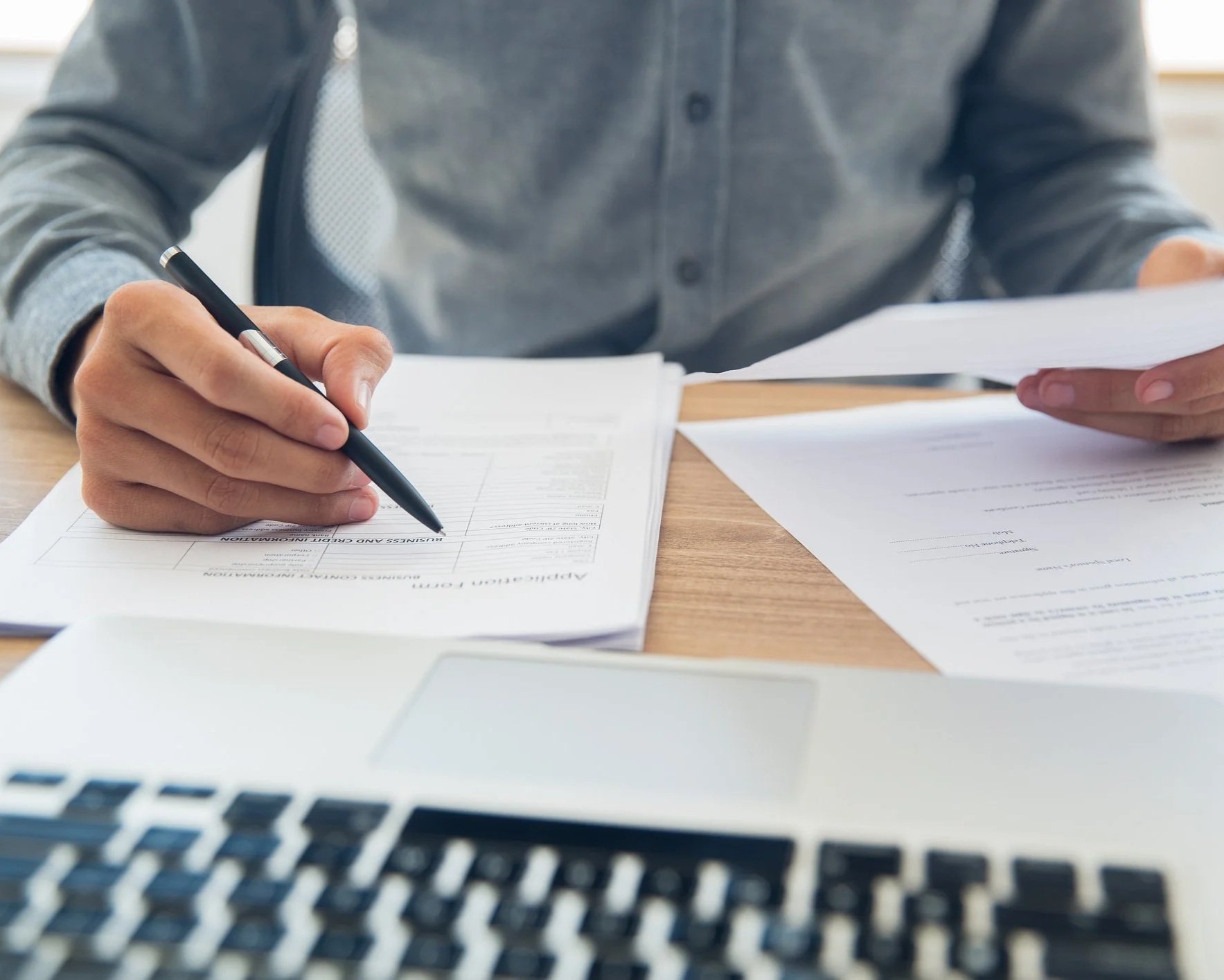 Person reviewing documents at a desk with a laptop, holding a pen and papers.