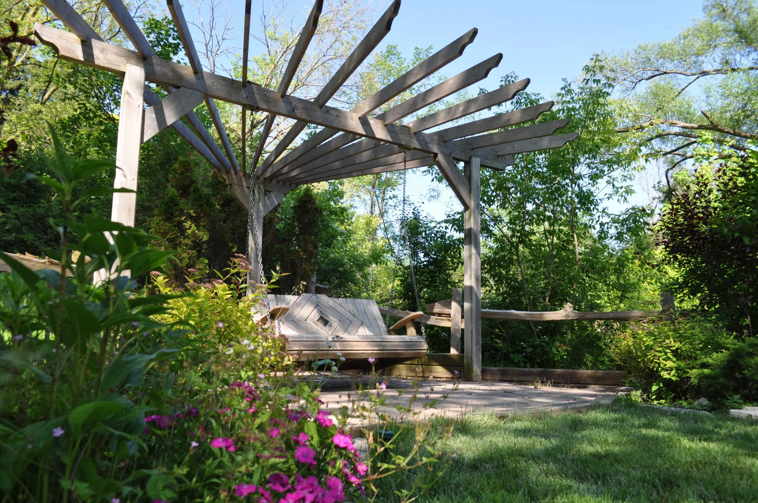 Wooden arbour with a hanging porch swing, surrounded by greenery and colorful flowers on a sunny day.
