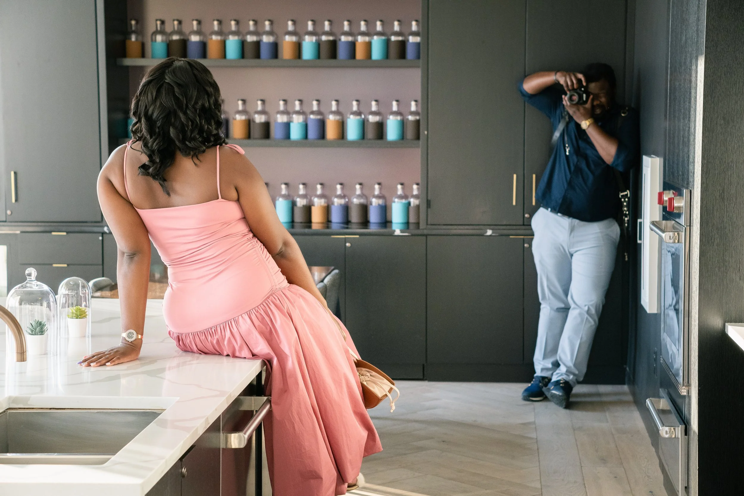 A woman in a pink dress posing on a kitchen counter while a photographer takes her picture in a modern kitchen with dark cabinetry and shelves of colorful jars.