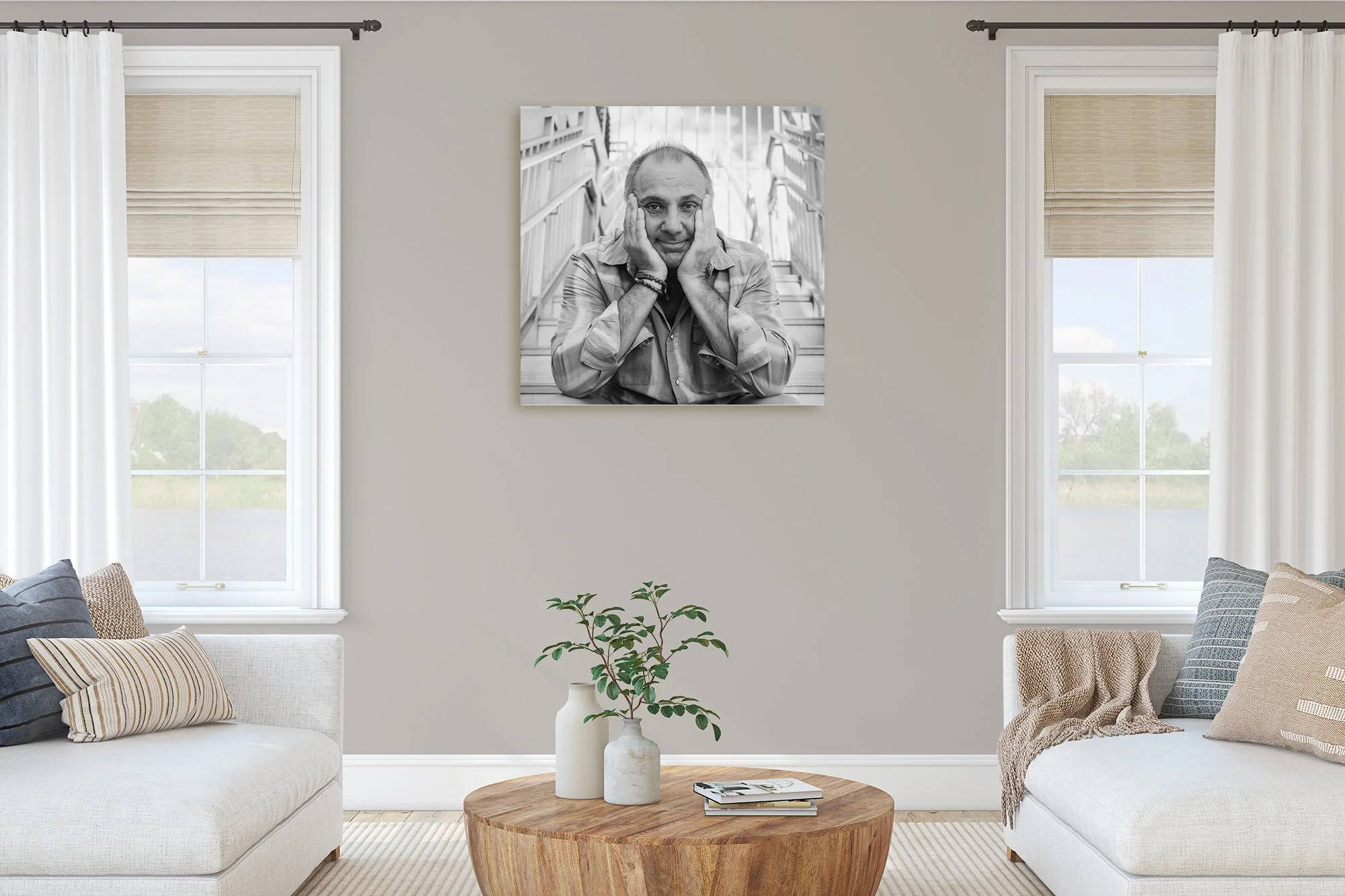 Living room with beige walls, featuring a black and white portrait on the wall, two windows with light-colored curtains, a round wooden coffee table with small plants, and a beige sectional sofa with striped and patterned pillows.