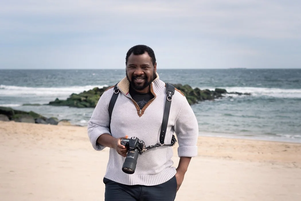 A man with a beard and short hair standing on a beach, smiling, holding a camera, with the ocean and rocks in the background.