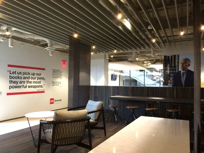 Modern indoor lounge with chairs, tables, and a bar area, featuring a CNN Diversity poster and a large portrait of a man in a suit on the wall, with exposed ceilings and hanging lights.