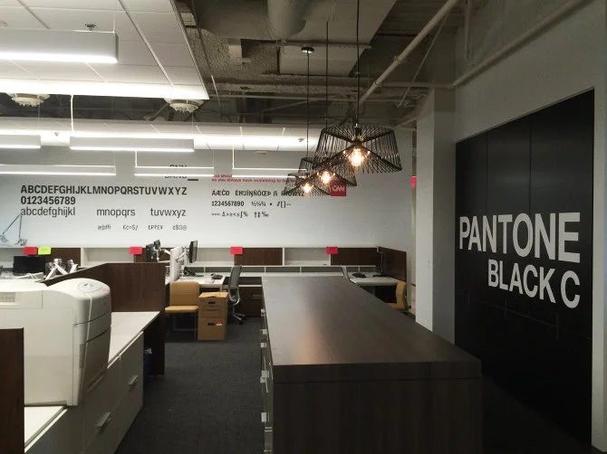 Office workspace featuring a large Pantone Black C sign on the wall, a long dark wood table, chairs, desks with computers, and a unique hanging light fixture. The ceiling is open with visible ductwork and fluorescent lighting.