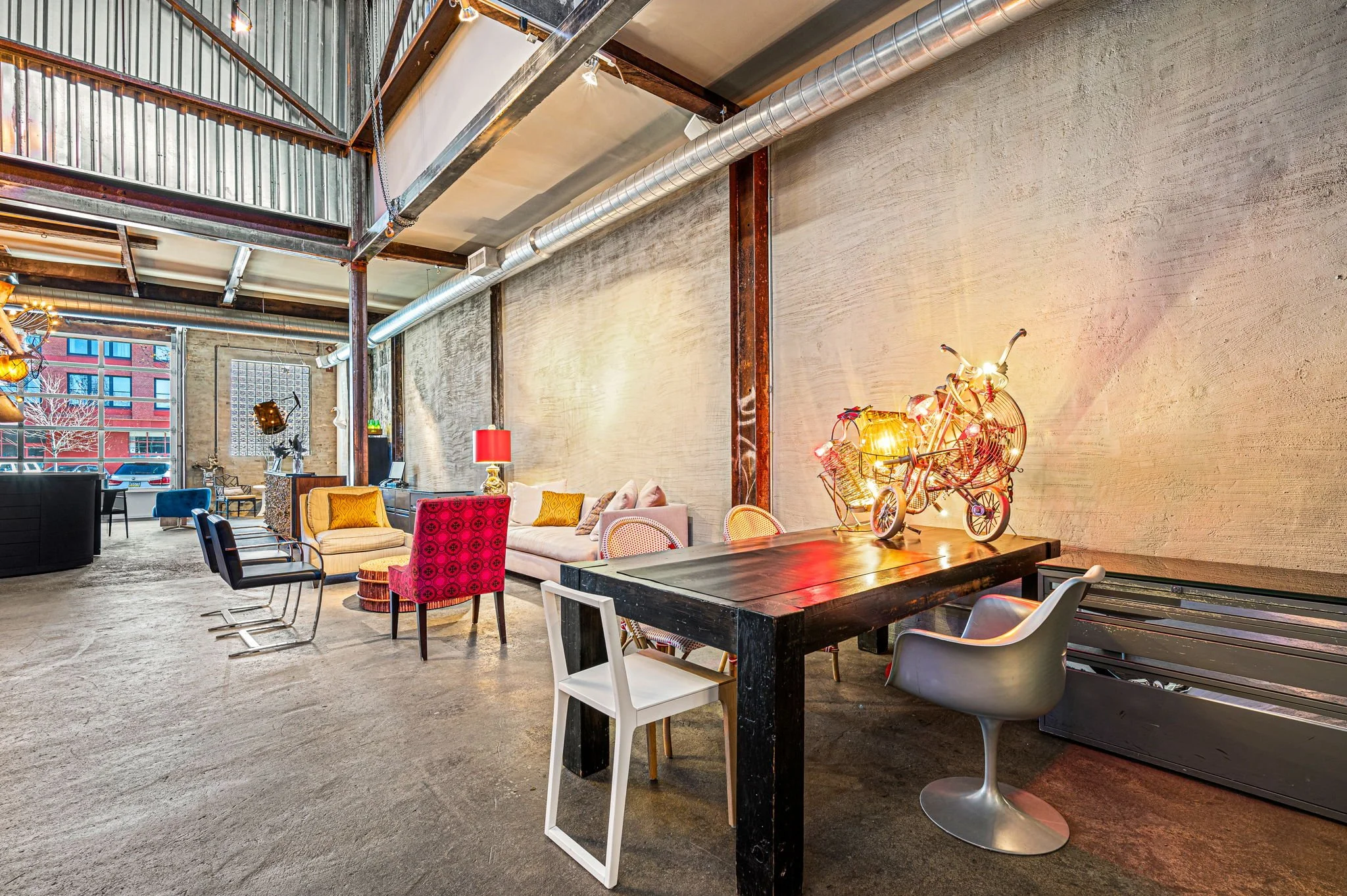 Interior of a modern, industrial-style living space with exposed concrete walls, colorful chairs, a sofa, a large black dining table with eclectic decor, including a bicycle-shaped lamp with lights, and a large window showing buildings outside.